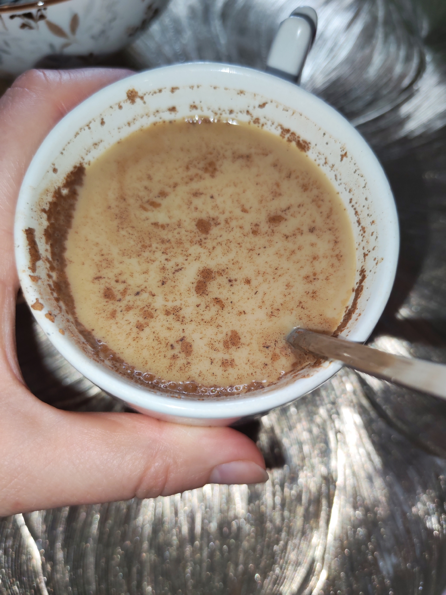 A hand holds a white ceramic cup filled with a light brown liquid, likely coffee or tea, with a sprinkling of brown powder, possibly cinnamon, on the surface and along the rim. A silver spoon is partially submerged in the liquid. The background appears to be a textured, metallic surface with a soft, blurred effect, suggesting a decorative placemat or tablecloth. The scene is lit by overhead lighting, creating highlights on the liquid and the metallic background. The image does not contain any information that would allow for localization to Izhevsk, Russia.
