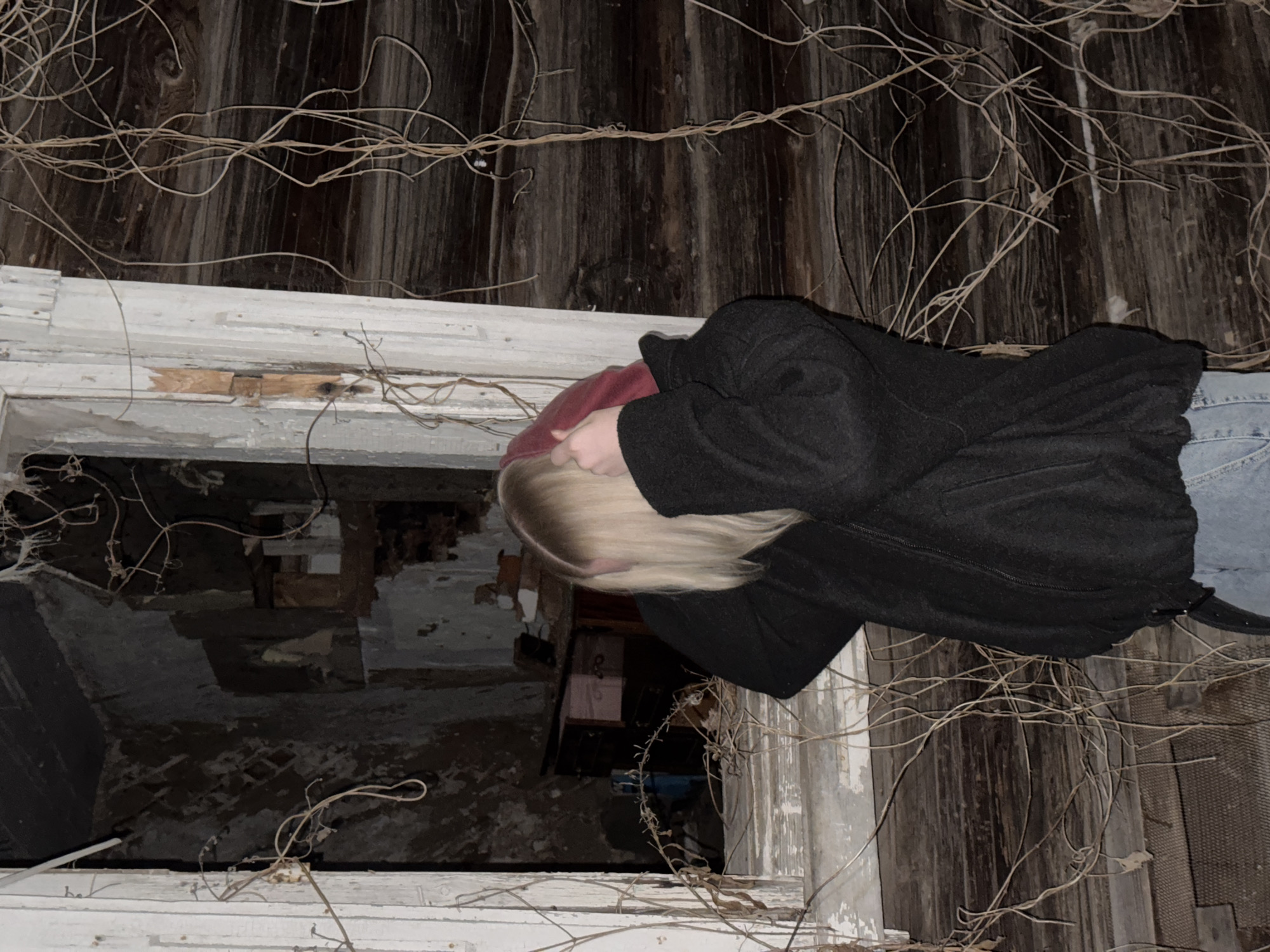 A person with blonde hair, wearing a dark coat, red hoodie, and jeans, stands at a window looking into a dilapidated interior. The building has weathered wooden walls covered with dry vines. The window frame is painted white and shows signs of deterioration. The interior seen through the window reveals a ruined space with debris and damaged walls. Zhigalki, Russia is not discernible in this image.