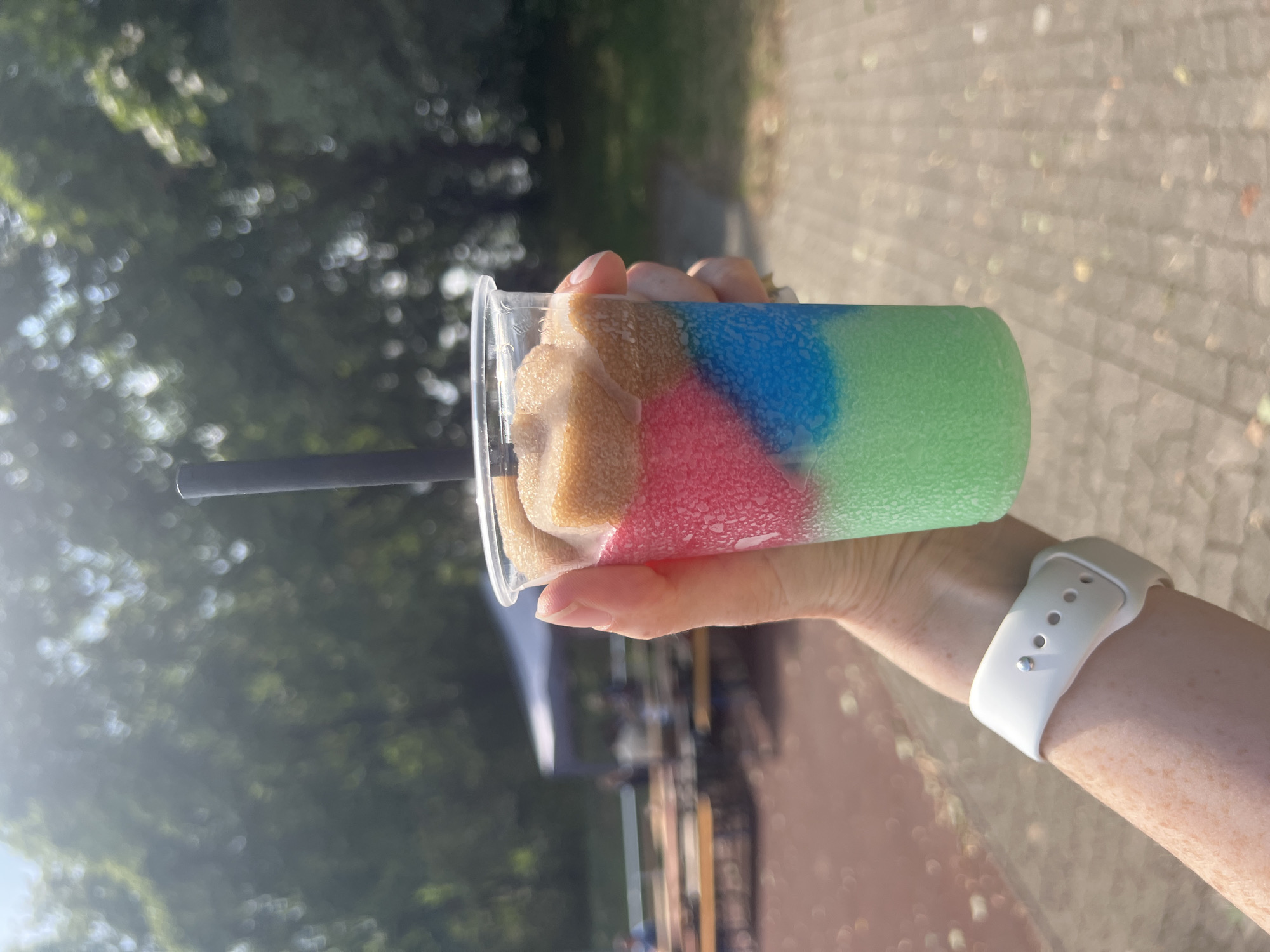 A hand, adorned with a white smartwatch on the left wrist and freckles, holds a clear plastic cup containing a layered, slushy drink. The drink has distinct layers of green, blue, pink, and a light brown or beige color on top, with a black straw inserted. The background is blurred and shows a sunny outdoor scene with trees, a white tent, picnic tables, and what appears to be a sports field or track. The setting suggests a casual outdoor event or park in Freising, Germany.