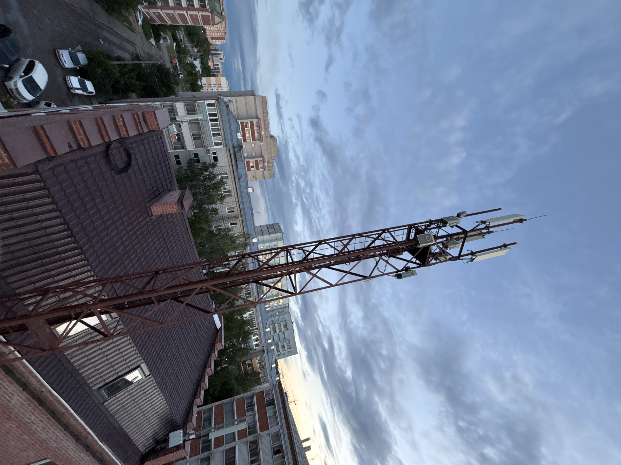 The image depicts a telecommunications tower situated on the roof of a building in Blagoveshchensk, Russia. The tower is constructed of red metal and is topped with multiple antennas. Below the tower, a dark brown tiled roof is visible, with several skylights. To the left of the tower, a street runs alongside apartment buildings. Several cars, including a white hatchback and two silver sedans, are parked on the street. The sky above is cloudy with a gradient of blue and grey, suggesting it is either early morning or late evening. The overall scene is an urban environment with a focus on infrastructure.