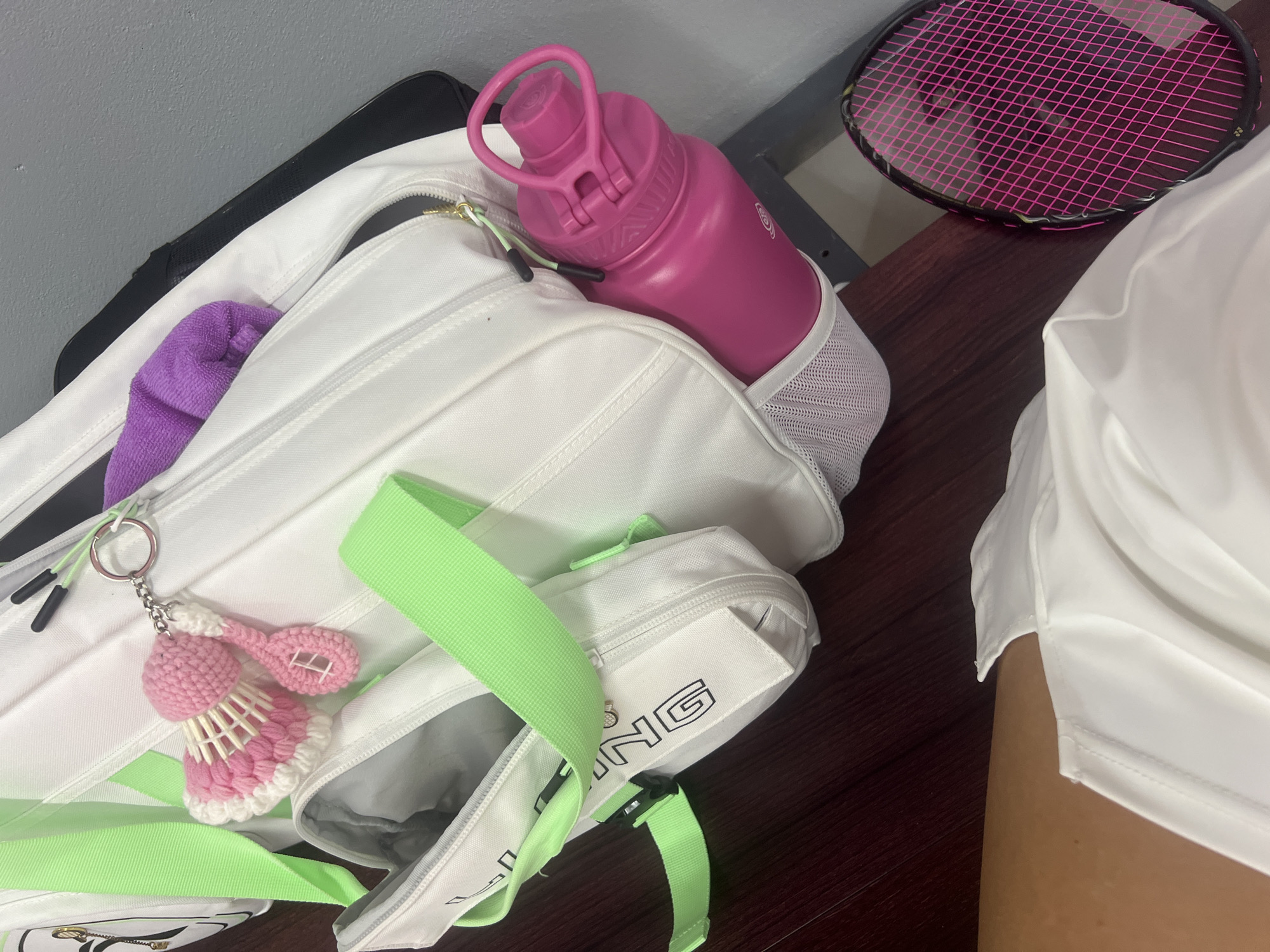 The image shows a partially filled white sports bag on a dark wooden surface. The bag contains a purple towel and a pink water bottle. A badminton racket is visible next to the bag. A crocheted badminton shuttlecock keychain hangs from the bag. A person wearing a white garment is partially visible on the right side of the frame. The background is a light gray wall.