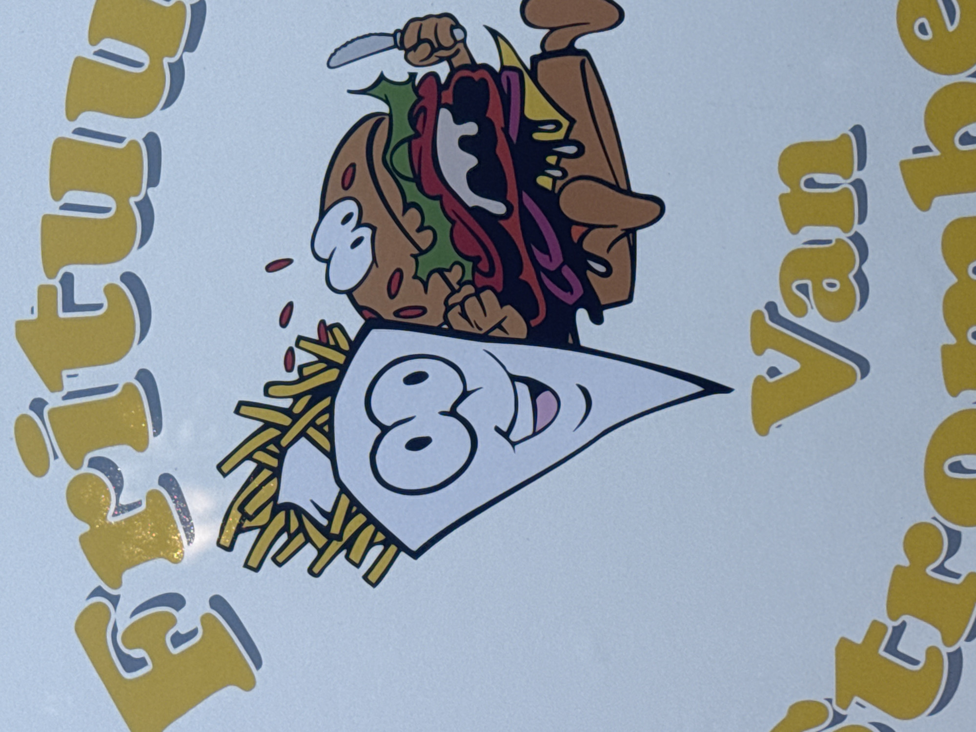 The image displays a cartoon graphic featuring a French fry character and a hamburger character running. The French fry character is white with black outlines and has large eyes and a smiling mouth, with French fries sticking out of its top. The hamburger character is brown with a bun, lettuce, tomato, cheese, and a dripping sauce, and it is holding a knife in its right hand. Both characters are depicted with legs and appear to be in motion. Above them, large yellow text with a grey shadow reads "Frituu", suggesting a name for a fast-food establishment. Below, smaller yellow text with a grey shadow reads "Van", and further below, partial yellow text with a grey shadow reads "trome be". This suggests the name of the establishment is likely "Frituu Van Trombe". The background is a plain white surface. The context provided indicates this image is located in Strombeek-Bever, Belgium, likely representing a local business or advertisement.