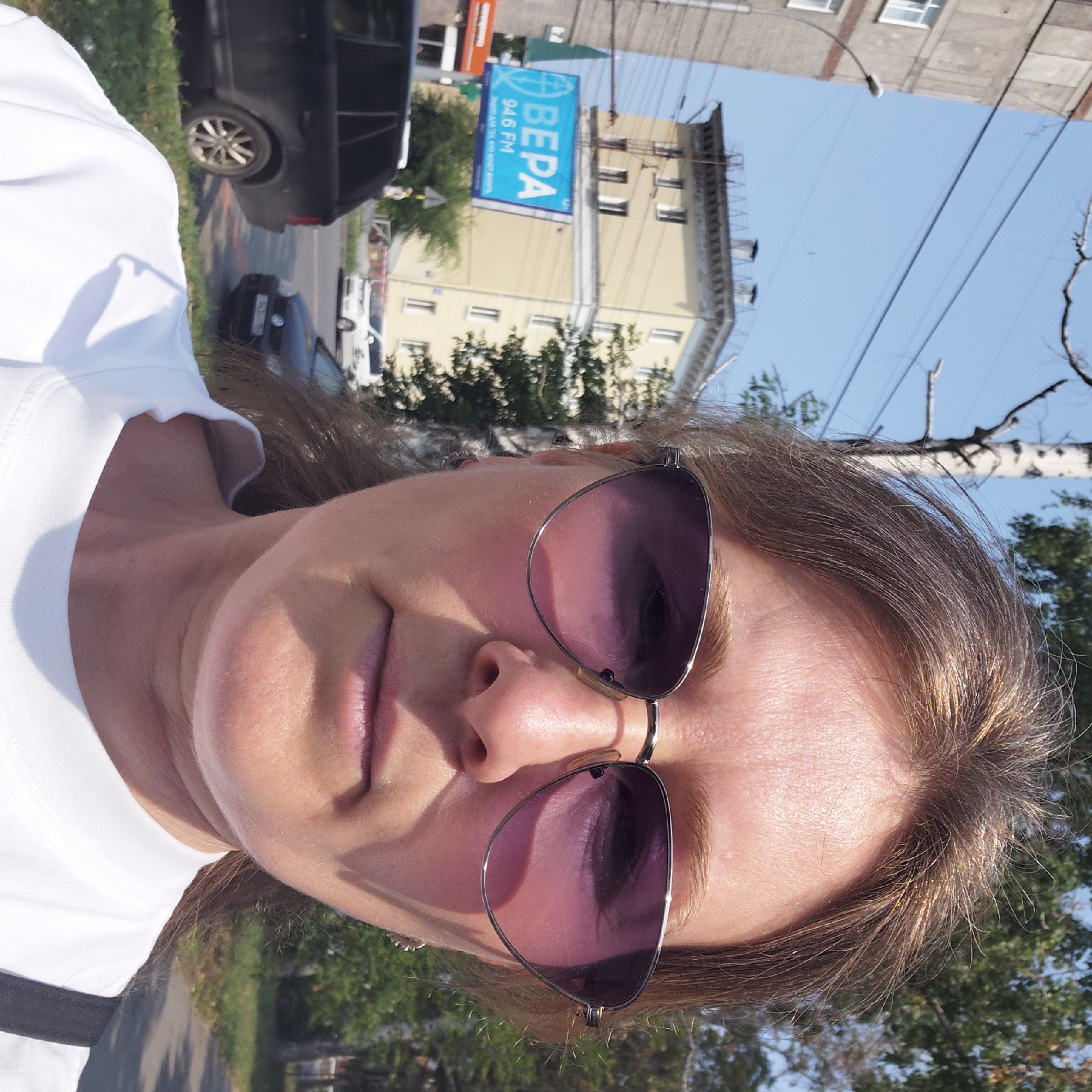The image is a selfie taken outdoors in a city. The photographer, a woman with brown hair and sunglasses, is in the foreground, her face partially obscured by the lenses. She is wearing a white t-shirt. In the background, there are buildings, trees, power lines, and cars. One building is a light yellow apartment complex with multiple windows. A blue billboard advertising "VERA 94.6 FM" is also visible. A dark SUV and a black car are parked on the side of the road. The sky is clear and blue. The scene appears to be a street in Novosibirsk, Russia, given the Cyrillic text on the billboard and the architectural style of the buildings, which is consistent with Soviet-era construction often found in Russian cities.