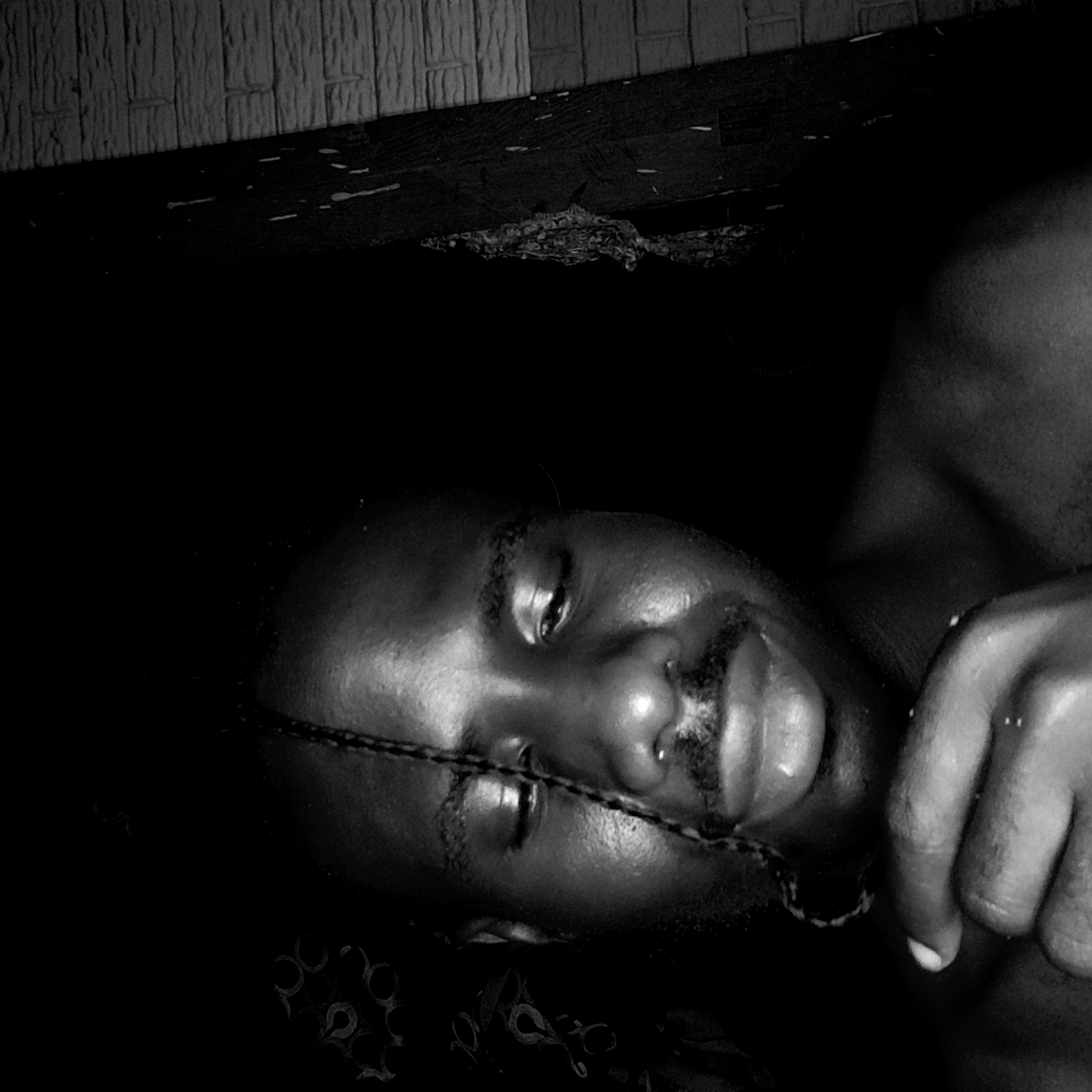 A black and white photograph depicts a person lying down. The person has dark skin, a thin braided cord across their face, and a slight smile. Their right hand is visible near their face. The background is dark, with a section of patterned fabric visible beneath the person and a wooden plank-like structure with markings above them. There are no indications of a location in Narayi, Nigeria.
