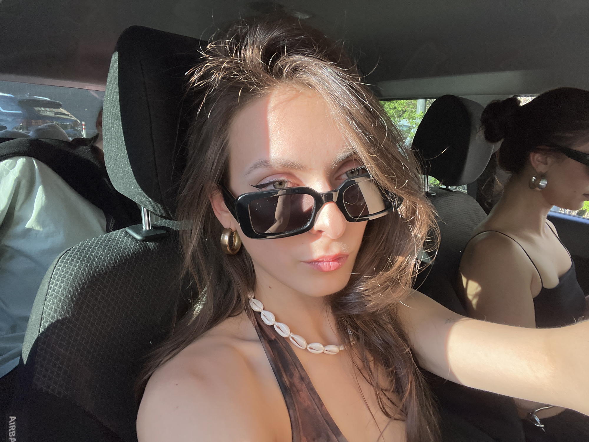 This image captures the interior of a vehicle, in Krasnoyarsk, Russia, on a sunlit day. A young woman, positioned in the front passenger seat, serves as the primary subject, looking directly towards the camera. She has long brown hair, styled with some strands appearing windswept or static, and is wearing large, dark, rectangular sunglasses. Her eyes are visible above the sunglasses, featuring prominent black winged eyeliner. She wears a brown, patterned halter-neck top and a distinctive necklace made of white cowrie shells. A single gold-toned hoop earring is visible on her left ear, while her right arm, extended out of frame to hold a device, is brightly illuminated by direct sunlight.
To her right and slightly behind her, a second female occupant is visible in profile, with dark hair tied back in a bun and wearing a black spaghetti-strap top and silver-toned hoop earrings. Further back on the left side, behind the front passenger seat, a third individual is partially visible, wearing light-colored clothing (possibly light green or white) and partially obscured by a dark headrest. The car's interior is dark, with textured fabric seats and an "AIRBAG" label visible on the side of the front passenger seat. Strong sunlight streams into the vehicle from the right, casting sharp shadows and creating bright highlights, particularly on the subjects' hair, skin, and the car's interior. Through the windows, green foliage is visible, and in the distant background, indistinct forms of other vehicles and structures can be discerned.