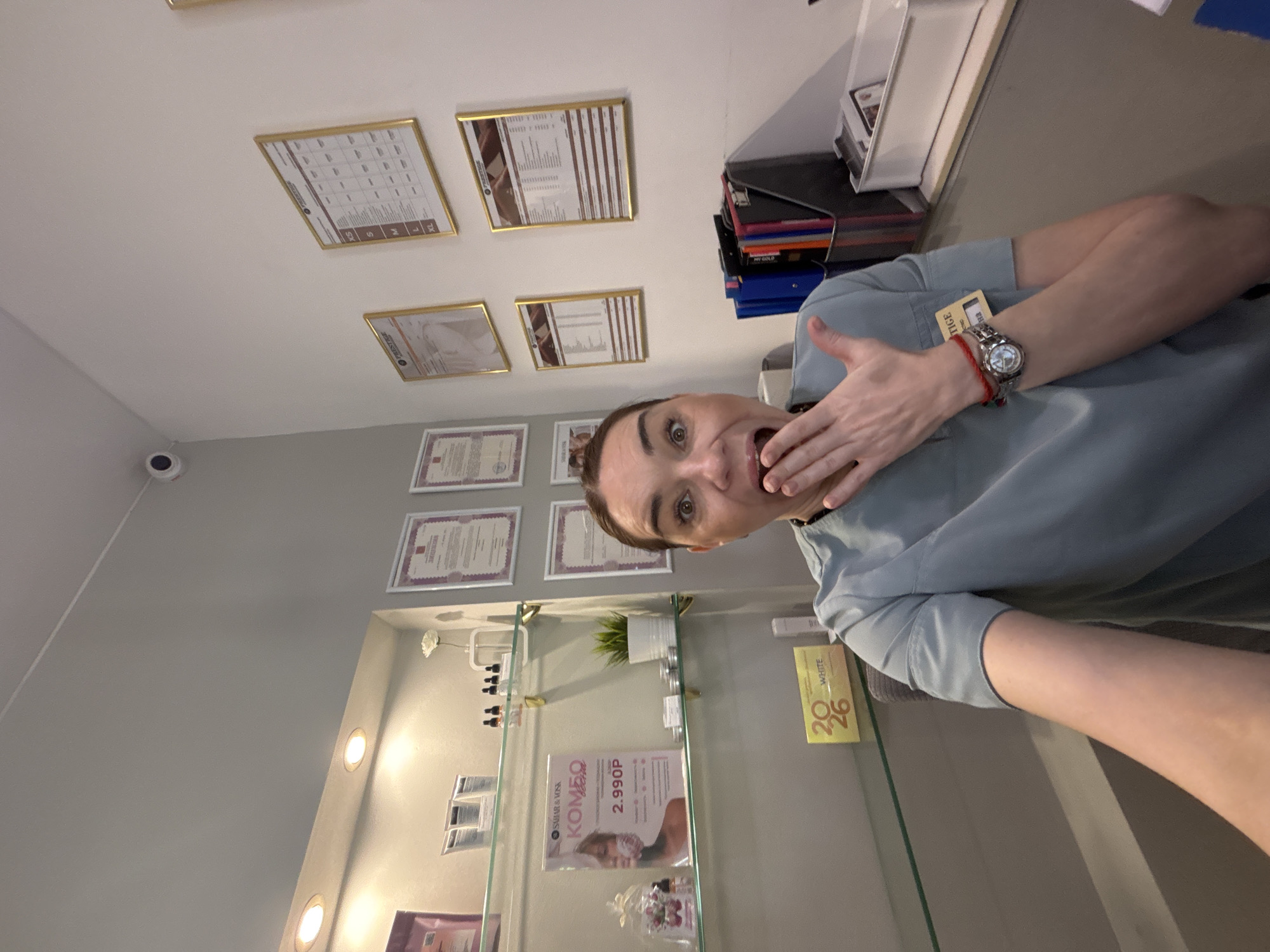 A person with brown hair and fair skin wearing a light blue uniform with a name tag on it is posed in a room with a glass shelf and several certificates or documents framed and hanging on the wall. The person has their hand raised to their mouth with an expression of surprise or shock, and a watch on their wrist. The glass shelf displays products, including one with the price "2.990P," and a box with the number "2026" on it. The room appears to be a medical or beauty clinic, characterized by professional displays and wall-mounted certificates. A security camera is visible on the ceiling. The location is Unknown, Unknown.
