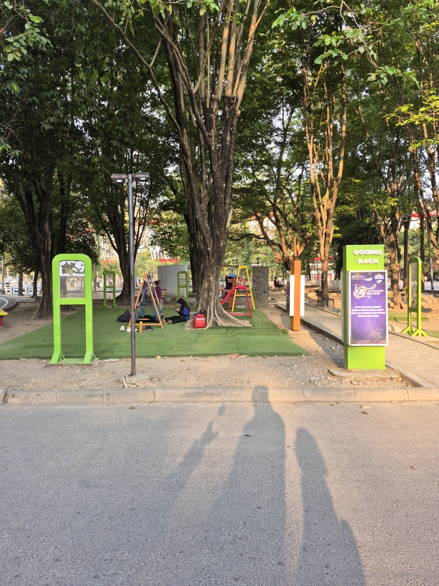 This is an exterior, medium-shot, eye-level, eye-level photograph of a public park in Surakarta, Indonesia. The park features artificial turf, exercise equipment, and trees. Two people are visible, with one sitting on a swing and another seated on the ground, looking at a phone. The photograph is illuminated by natural sunlight, casting long shadows of two individuals onto the asphalt road in the foreground.