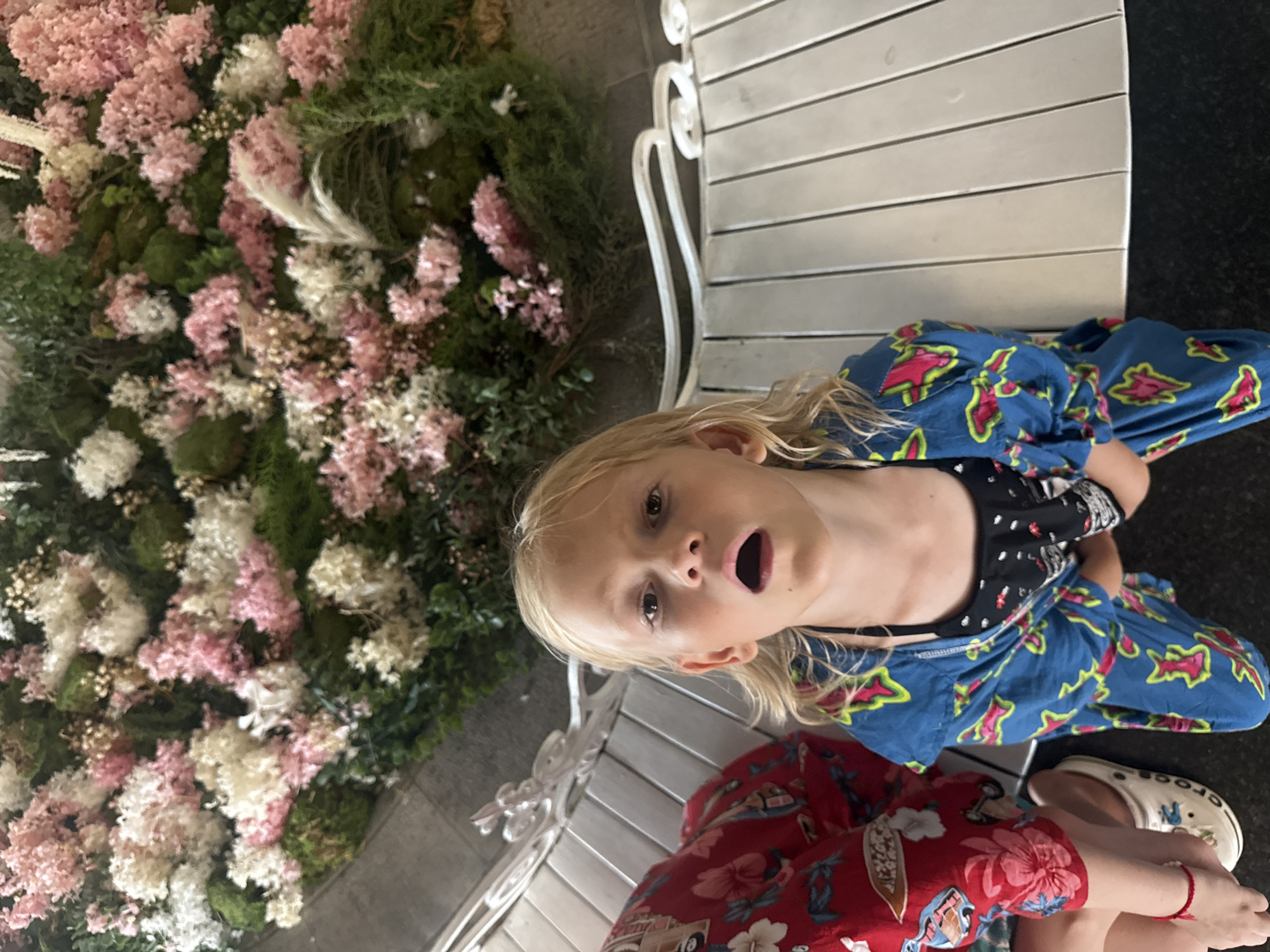 In the city of Yalta, Ukraine, a young girl with blonde hair, wearing a blue patterned robe over a black and white top, sits on a white slatted bench. Her mouth is open in surprise or excitement. To her left, another person, only their arm and shoulder visible, wears a red shirt with a floral pattern. Both are situated in front of a vibrant display of pink and white flowers interspersed with greenery. The overall scene appears to be outdoors.