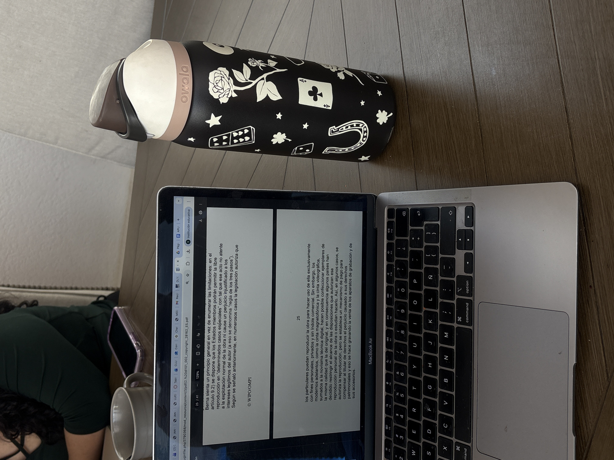 The image captures an indoor scene, likely in Puebla, Mexico, showing a person studying or working on a laptop. A black Owala brand water bottle with decorative white designs, including roses, stars, playing card symbols (Ace of Clubs prominent), dice, horseshoes, and four-leaf clovers, lies beside a silver MacBook Air laptop. The water bottle's lid band also features the "owala" logo.
The laptop screen displays a PDF document in Spanish, discussing copyright law and limitations, specifically referencing Article 9.2, the "regla de los tres pasos" (three-step test) regarding reproduction rights. The text mentions "WIPO/OMPI" and discusses personal, private, non-commercial use, as well as the impact of modern digital technology on reproduction and mechanisms for compensating rights holders. The document is page 25 of 41 and is zoomed to 129%. Several browser tabs are visible, including "Institución educativa," "APU," and "DLT."
Partially visible on the left is a person with dark, curly hair wearing a dark green top, along with a white mug and a smartphone in a clear case. The objects are placed on a light-colored wooden floor.
