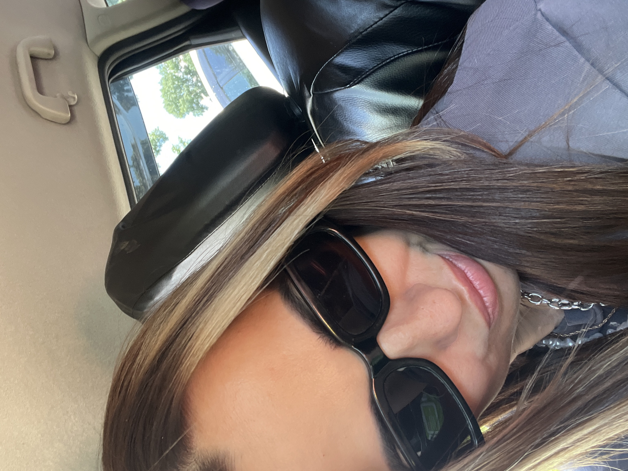 A close-up, indoor, selfie-style shot shows a woman with long, brown hair with blonde highlights, wearing black sunglasses and a grey shirt, and a silver chain necklace. She is seated in the passenger seat of a car, with the headrest of the driver's seat visible behind her. The car's interior is dark, with a glimpse of the outdoors visible through the window.