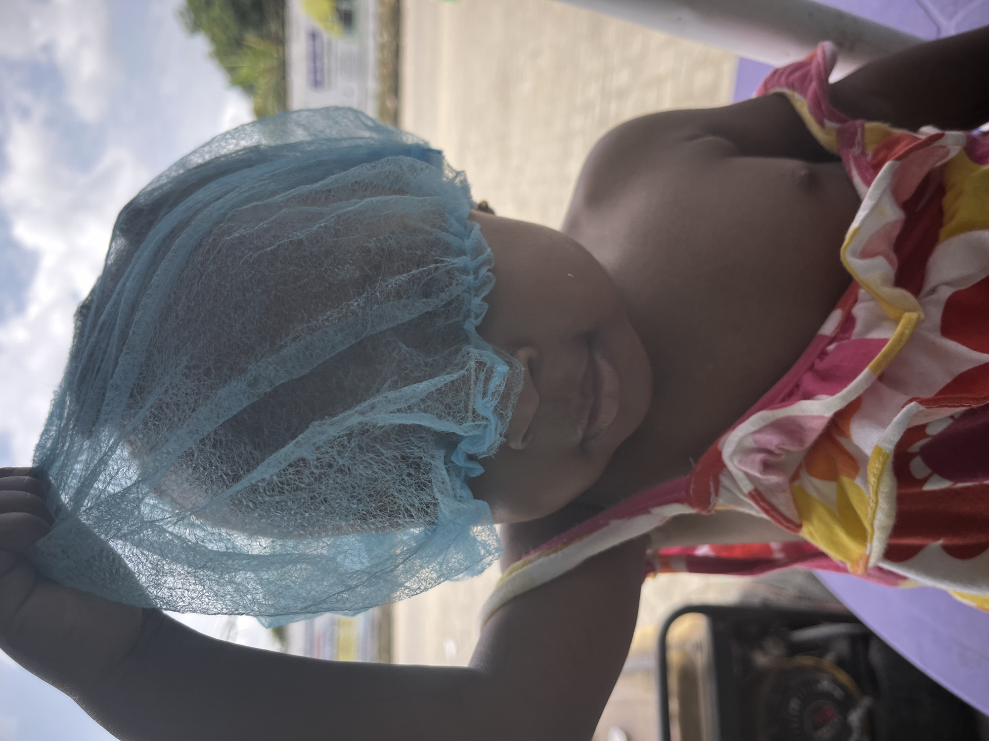 A close-up, angled shot shows a child wearing a light blue disposable hairnet. The child is dark-skinned and wears a patterned top with red, pink, white, and yellow colors. The child is seated, and the background reveals an outdoor setting with a sky that appears overcast. In the blurred background, there are hints of trees and a sign. The location appears to be Lekki, Nigeria, based on the surroundings and information provided.