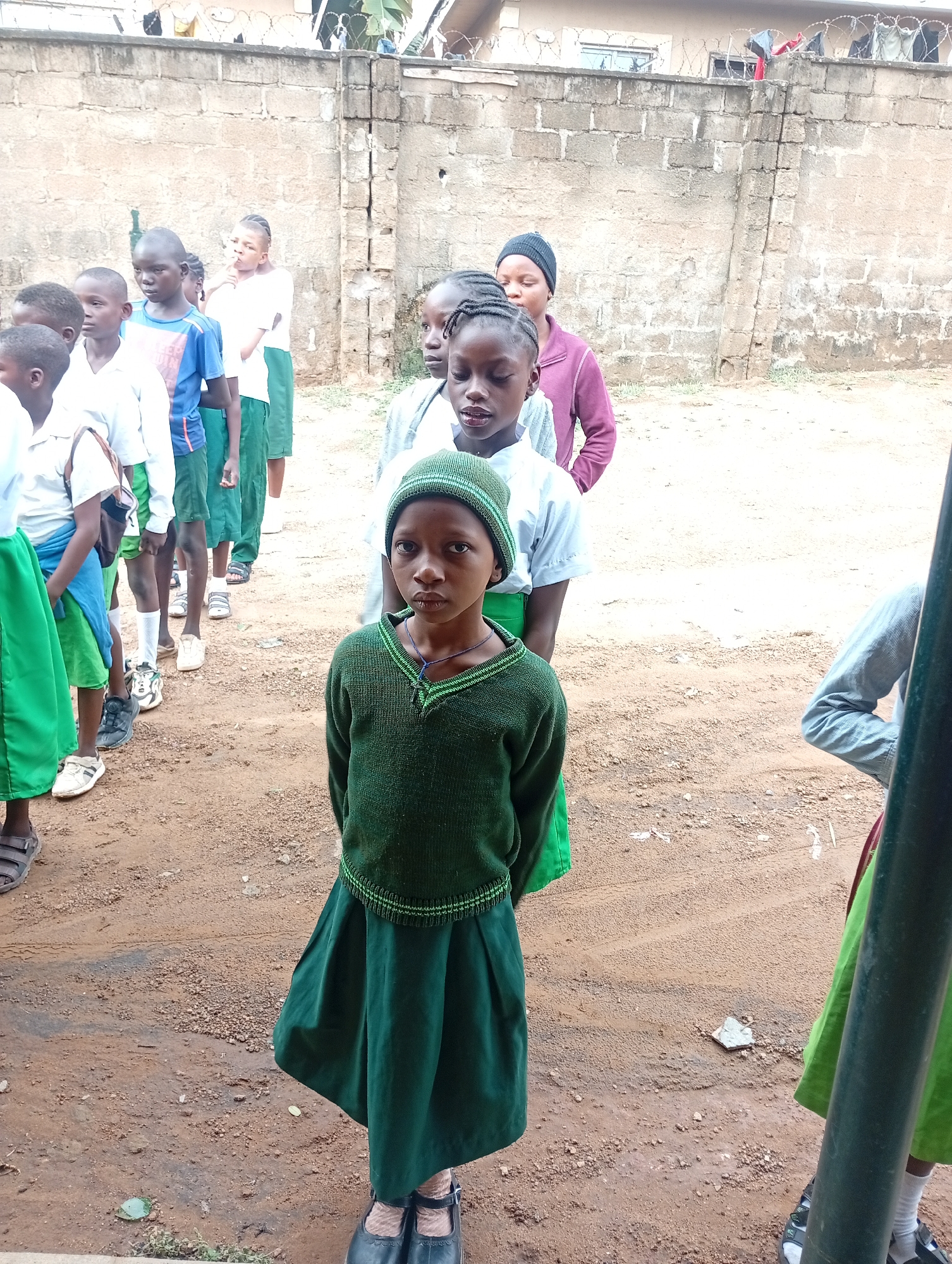 In Narayi, Nigeria, a group of children in school uniforms stand on a dirt ground, likely outdoors. The uniforms consist of green skirts or shorts, and some wear white shirts. A girl in the foreground wears a green sweater and matching hat. Behind her, the other children are lined up, some wearing beanies or head coverings. A brick wall forms a backdrop to the scene.