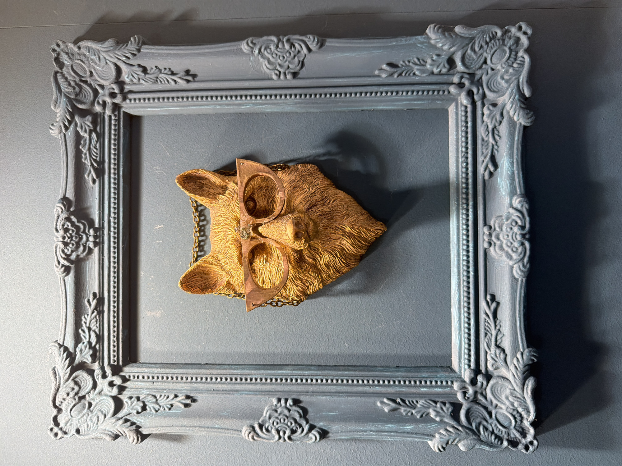 A decorative, ornate picture frame with a dark grey, distressed finish hangs on a textured, dark grey wall. Inside the frame, a golden fox head sculpture wearing round, copper-colored glasses is suspended by a chain. The fox sculpture has detailed fur texture and a slightly tilted head. The frame itself features elaborate scrollwork and floral motifs on its corners and sides.