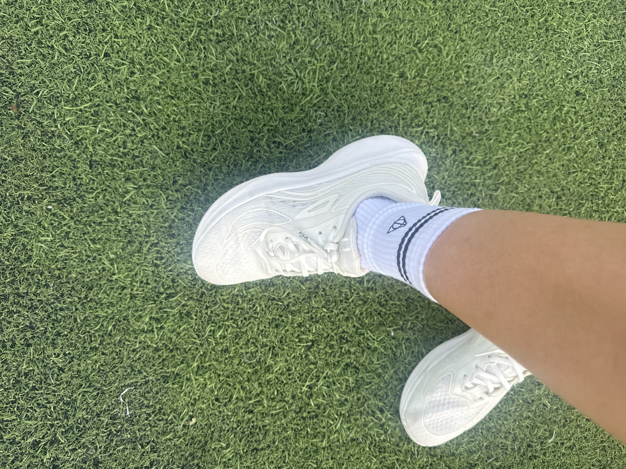 A person's legs are shown wearing white sneakers and white socks with black stripes on an artificial turf field in Baan Mae Nam, Thailand. One leg is bent at the knee, with the foot pointing upwards and to the right. The other leg is extended to the left, with the foot flat on the turf. The artificial turf is green and appears to be well-maintained. The lighting suggests it is daytime.