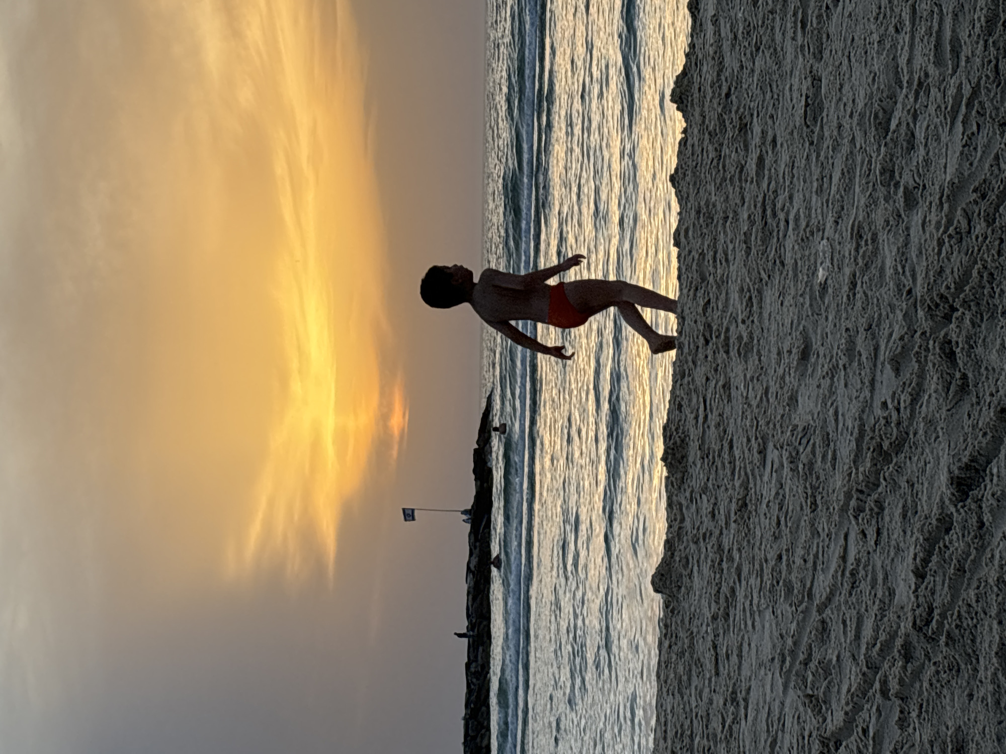 A young person stands in a swimsuit on the edge of a beach in Tel-Aviv, Israel, at sunset. The sea stretches to the horizon where it meets the sky colored with hues of orange and yellow. A rocky breakwater extends along the shoreline, marked by a small Israeli flag. The texture of the sand is visible on the shore.