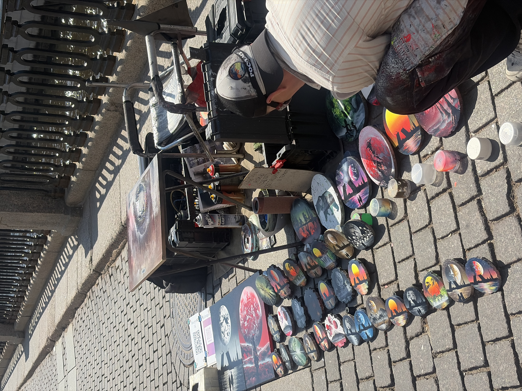 An artist is kneeling on a cobblestone street in Saint Petersburg, Russia, with a display of his painted wooden art pieces spread out before him. The artist, wearing a cap and a striped shirt, is looking down at the artworks, which are mostly oval-shaped and feature vibrant, abstract designs, some depicting landscapes with sunsets and trees. Spray paint cans and lids are scattered around the display. In the background, a metal fence lines a body of water, casting shadows on the street. There are also large black containers and a folding table with a painting on it. The scene suggests an outdoor market or street art exhibition.