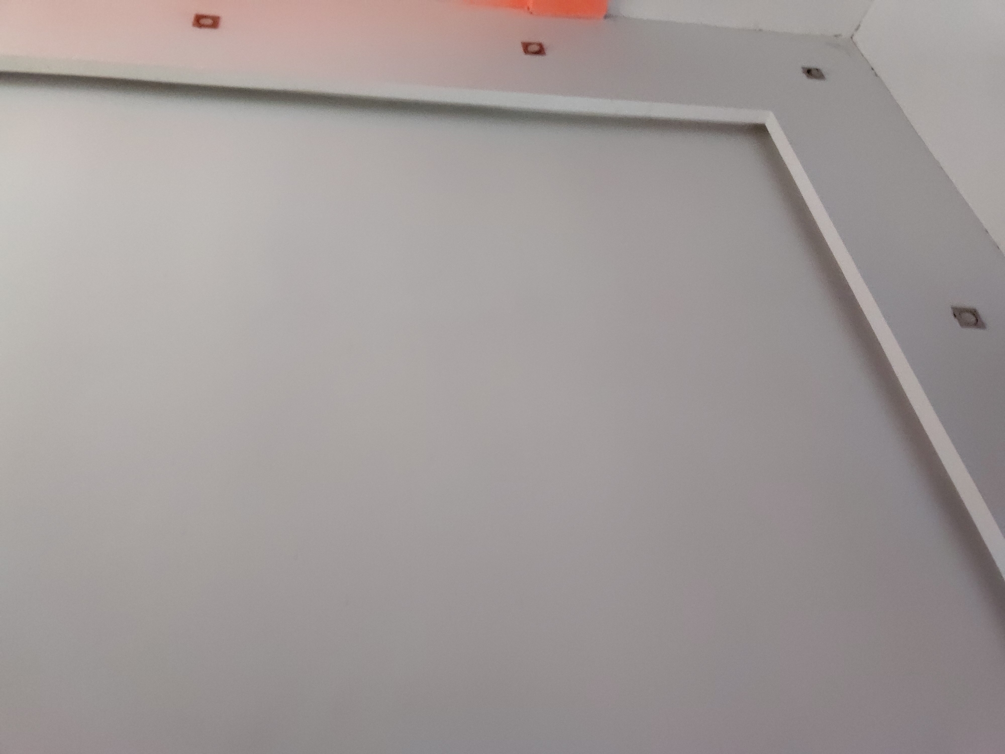 The media file presents an interior view of a room, likely undergoing construction, renovation, or recently completed and unfurnished. The dominant feature is a large, unadorned light gray wall, which fills most of the frame.
A prominent white rectangular frame or molding is affixed to the wall, creating a clean, defined border around a central, slightly recessed or raised panel area, also in light gray. This architectural detail suggests a designated space, possibly for a television or a decorative element.
Along the upper edge of the wall and ceiling, several small, dark, square or circular openings are visible. These appear to be cutouts for recessed lighting fixtures or electrical outlets that have not yet been installed. Towards the top-center of the image, an area of bright orange material or structure is visible on the ceiling, possibly exposed conduit, insulation, or an unfinished ceiling panel, reinforcing the impression of ongoing work.
The color palette is predominantly neutral, featuring shades of light gray and white, with the contrasting orange detail. The lighting appears soft and uniform, without strong shadows, indicating either ample ambient light or diffused artificial illumination. No human activity is depicted, and there is no visible text. The image provides a close-up perspective of the structural and finishing details of the space in Santa Ana, Venezuela.