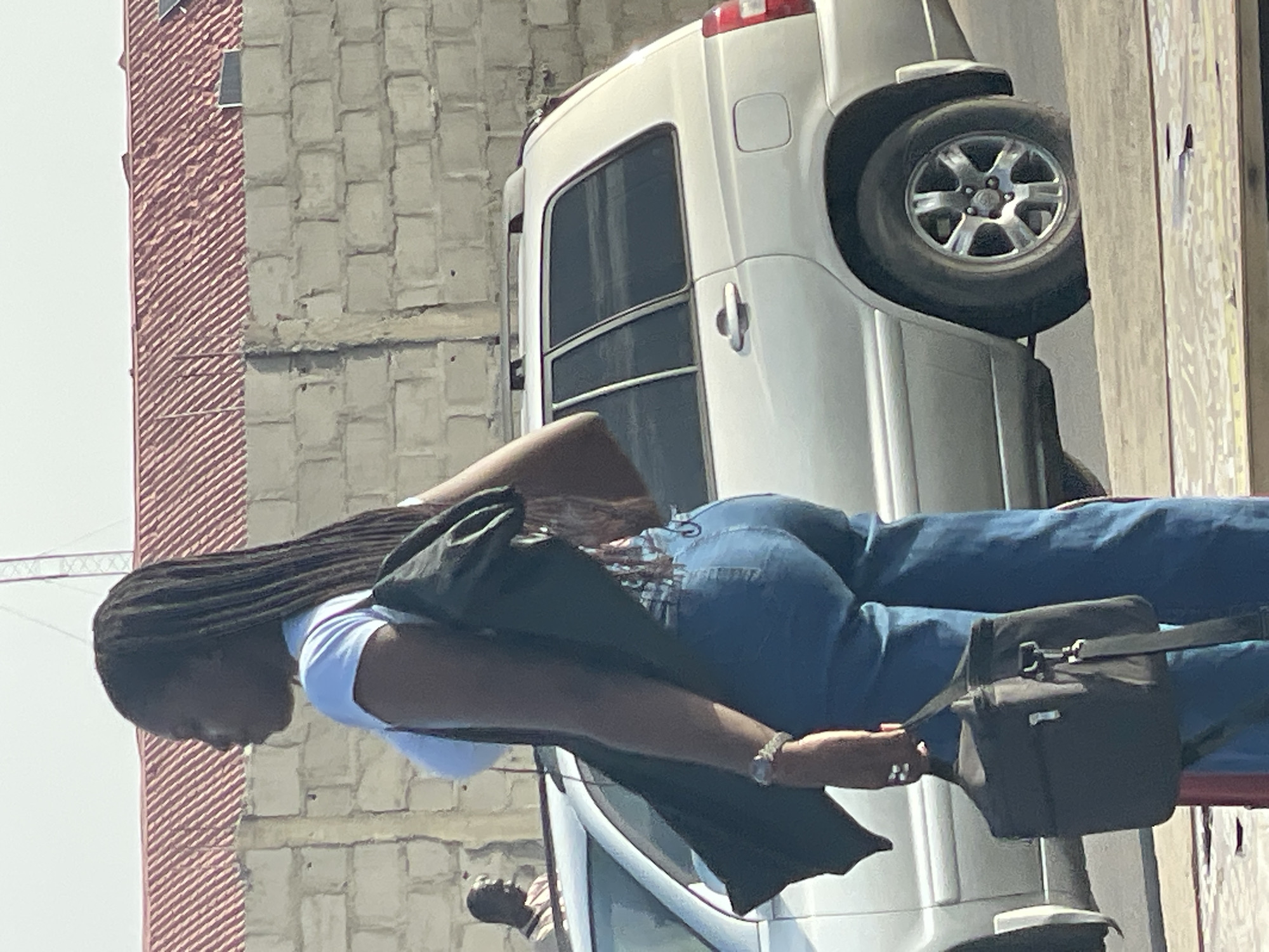 A medium shot depicts a black woman with long braids walking past a white SUV in what appears to be a city street. She wears a light blue top and blue jeans, a dark colored jacket and a small black bag hung over her shoulder. The background includes a grey concrete block wall and a brick structure. The photo was likely taken in the city of Ajara, Nigeria.
