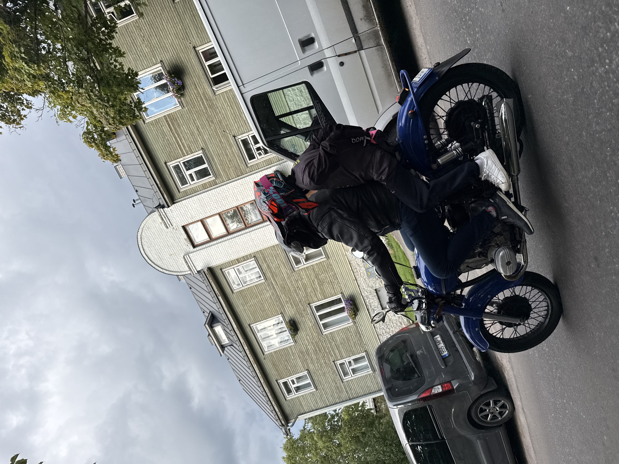 The image depicts a blue motorcycle with two individuals wearing helmets, riding down a street in Tallinn, Estonia. The rider in front is wearing a black leather jacket and dark pants, while the passenger behind is wearing a black jacket and dark pants. Both are wearing full-face helmets, with the passenger's helmet featuring a distinctive colorful design. The motorcycle appears to be older, with a visible engine and spoke wheels.
The background includes a two-story wooden building painted green, with several windows. A silver van is parked to the right of the motorcycle, and a dark gray van is visible to the left. The sky is overcast with visible clouds, suggesting a cloudy day. The street surface is asphalt. A license plate on the dark gray van reads "955 MPY". The architecture of the buildings and the style of the vehicles suggest an urban setting in Eastern Europe.