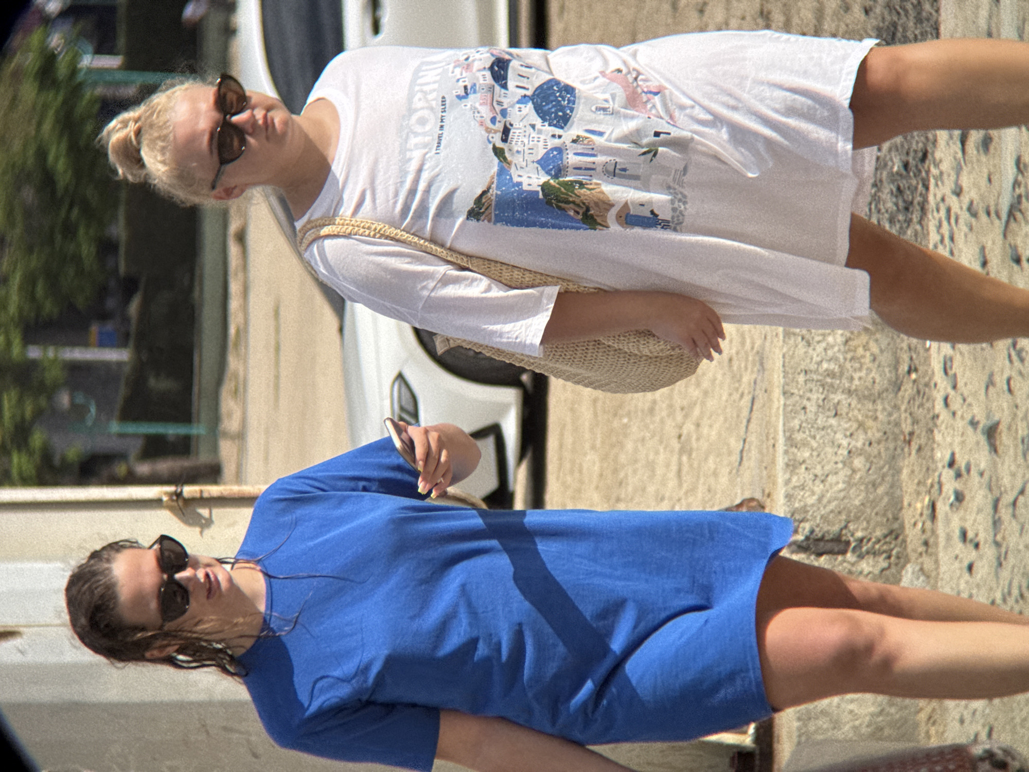 Two women are walking outdoors, one in a blue dress and the other in a white t-shirt dress. The woman on the left is wearing sunglasses and holding a phone. She has wet hair pulled back. The woman on the right is wearing sunglasses and has her hair in a bun. She is carrying a woven bag over her shoulder. Both women are on a paved walkway with a sandy area to their right. In the background, there is a white car and some greenery. The scene appears to be in a sunny outdoor location, possibly a resort or tourist area. The context provided about Nha Trang, Vietnam is not visible in the image itself.