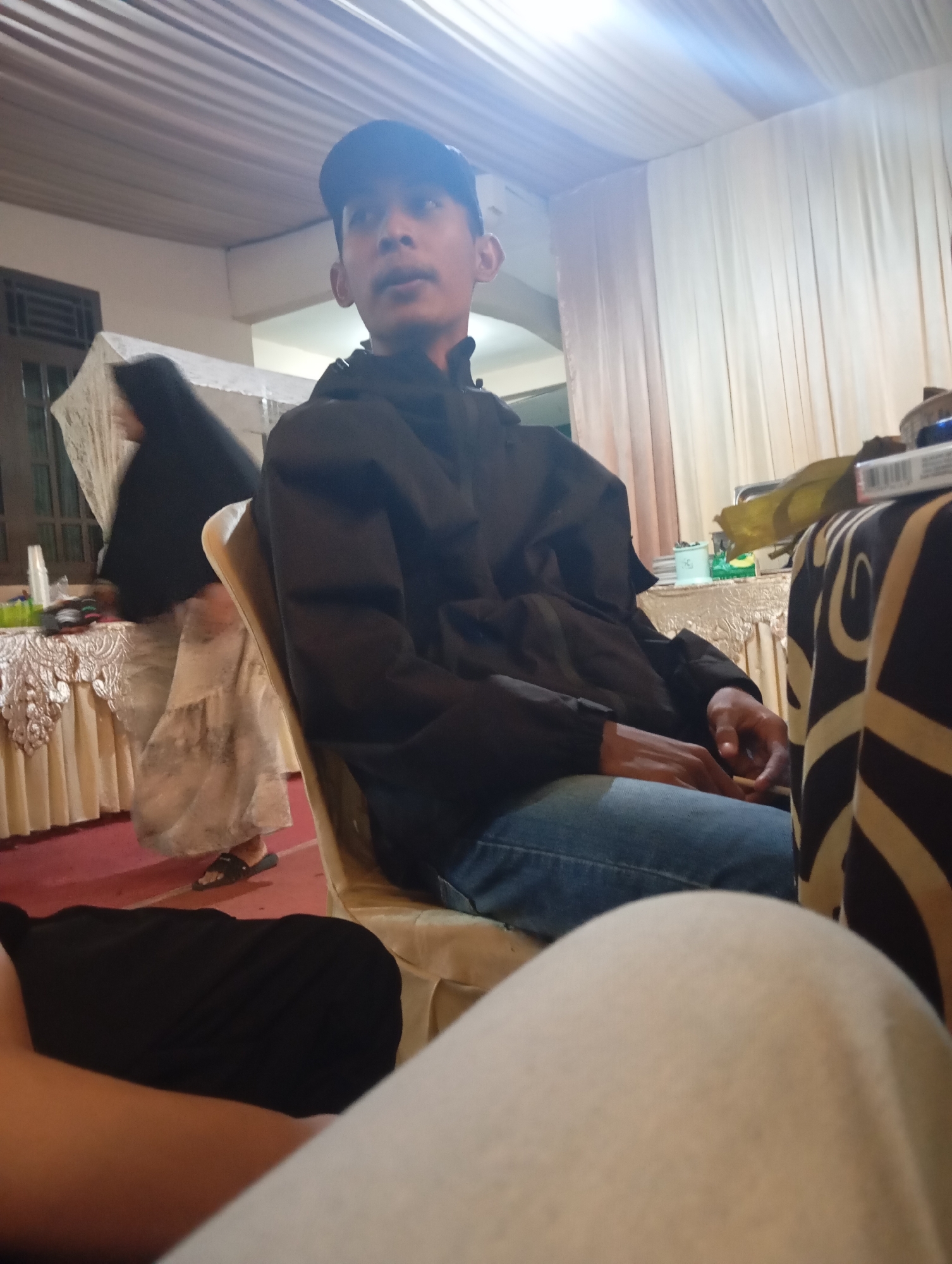 The image depicts a low-angle shot of a young man wearing a black baseball cap and a black jacket, seated in a chair. He is looking upwards and to his left. In the background, a woman in a black hijab and a light-colored patterned dress is visible, standing near a table covered with a white lace tablecloth. The environment appears to be indoors, with white drapery hanging on the wall behind the man and a ceiling with visible beams and fabric covering. The overall lighting suggests an interior space, possibly decorated for an event. The location is identified as Kemangkon, Indonesia.