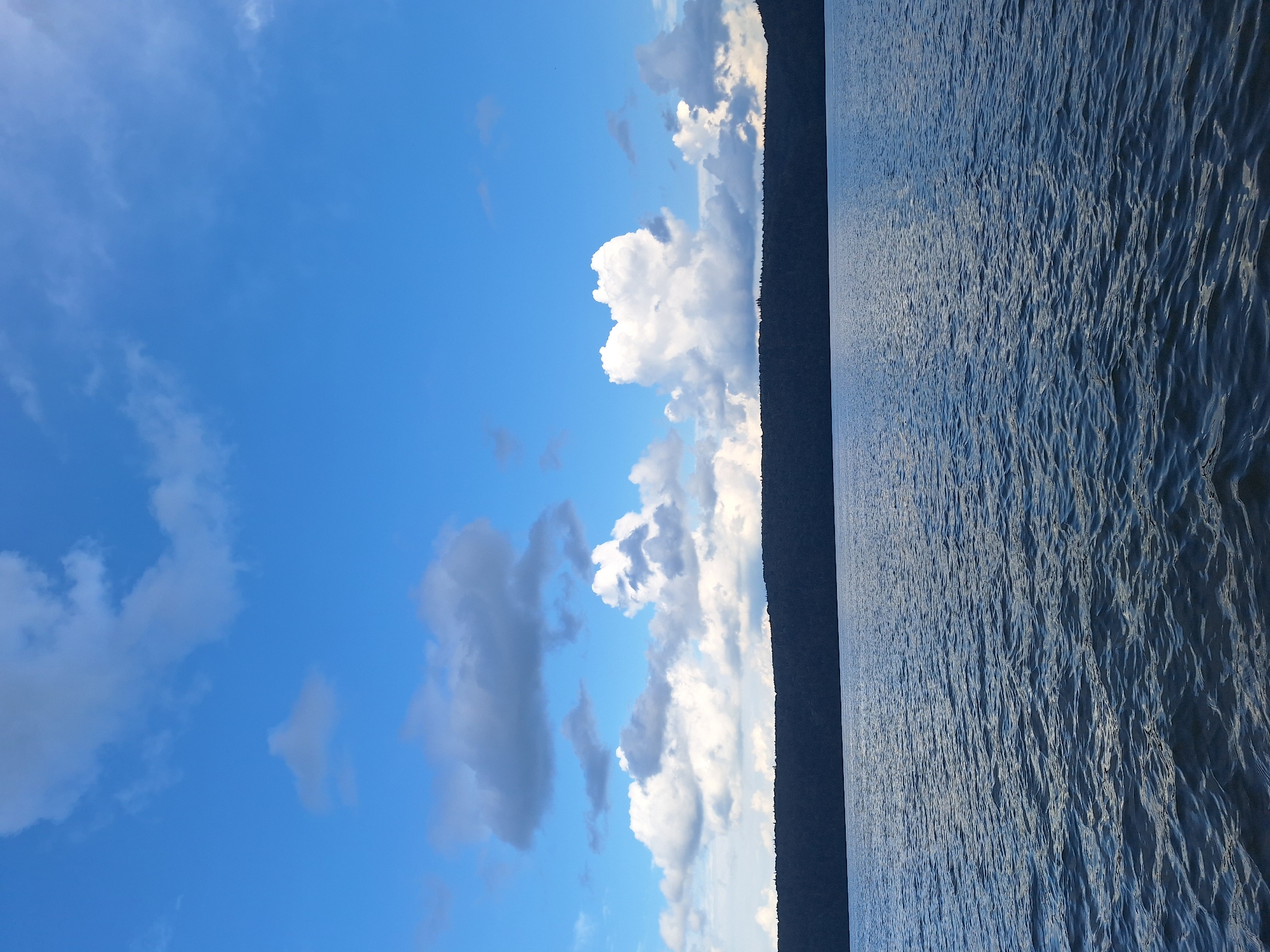The image depicts a body of water with a dark, tree-lined horizon under a blue sky with white and gray clouds. The water has small waves, indicating some movement. The scene appears to be natural and serene, with no visible signs of human activity or structures. The location is identified as the city of Атамановка, Russia.
