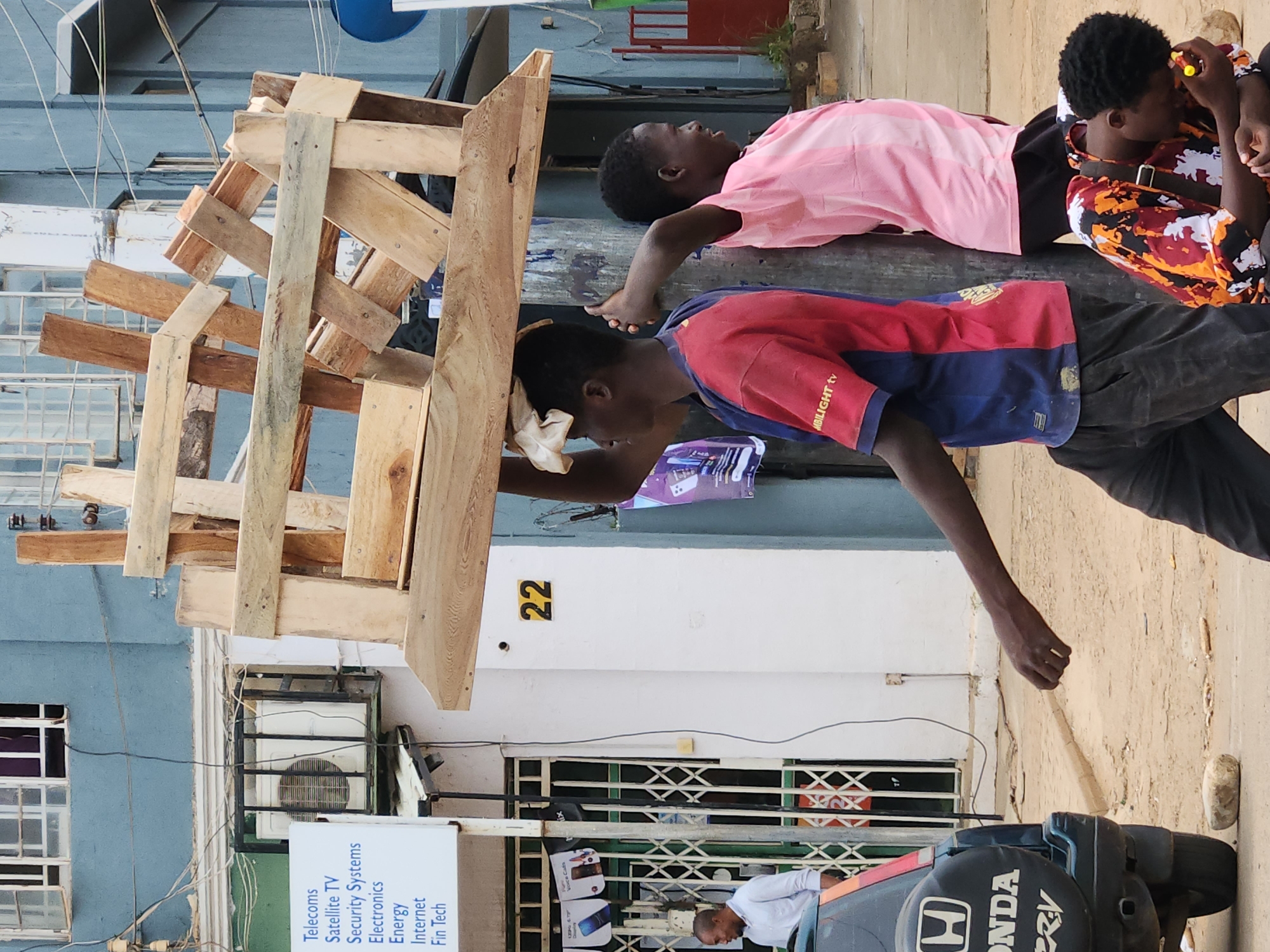 This media file captures an outdoor street scene, likely in an urban or commercial area, featuring several individuals and various functional objects.
In the center of the frame, three young males are resting on a large, dark, horizontal beam or pole. One boy, wearing a pink shirt, is lying on his back, looking upwards. Next to him, another boy in a red and blue shirt leans against the pole, looking downwards, with the text "MBILIGHT tv" visible on his shirt. A third boy, dressed in a multi-colored patterned shirt (orange, black, white), is sitting or leaning further along the pole, holding a small yellow object. In the lower right corner, a man in a white shirt is partially visible, bending over a black Honda generator, which bears the "H HONDA CR-V" logo.
A prominent, somewhat unstable-looking stack of wooden planks or palettes occupies the left side of the foreground. The background consists of buildings with light blue and white painted walls, featuring windows with metal security grates. An outdoor air conditioning unit is mounted on one of the walls. Numerous electrical wires are strung across the scene. A blue satellite dish can be seen in the upper background.
Notable details include a white sign listing various services: "Telecoms," "Satellite TV," "Security Systems," "Electronics," "Energy," "Internet," and "Fin Tech." The number "22" is painted in yellow on a white section of a wall. The overall impression is a candid moment of daily life, with the boys appearing relaxed or resting, possibly taking a break from activities in a bustling environment during daytime.