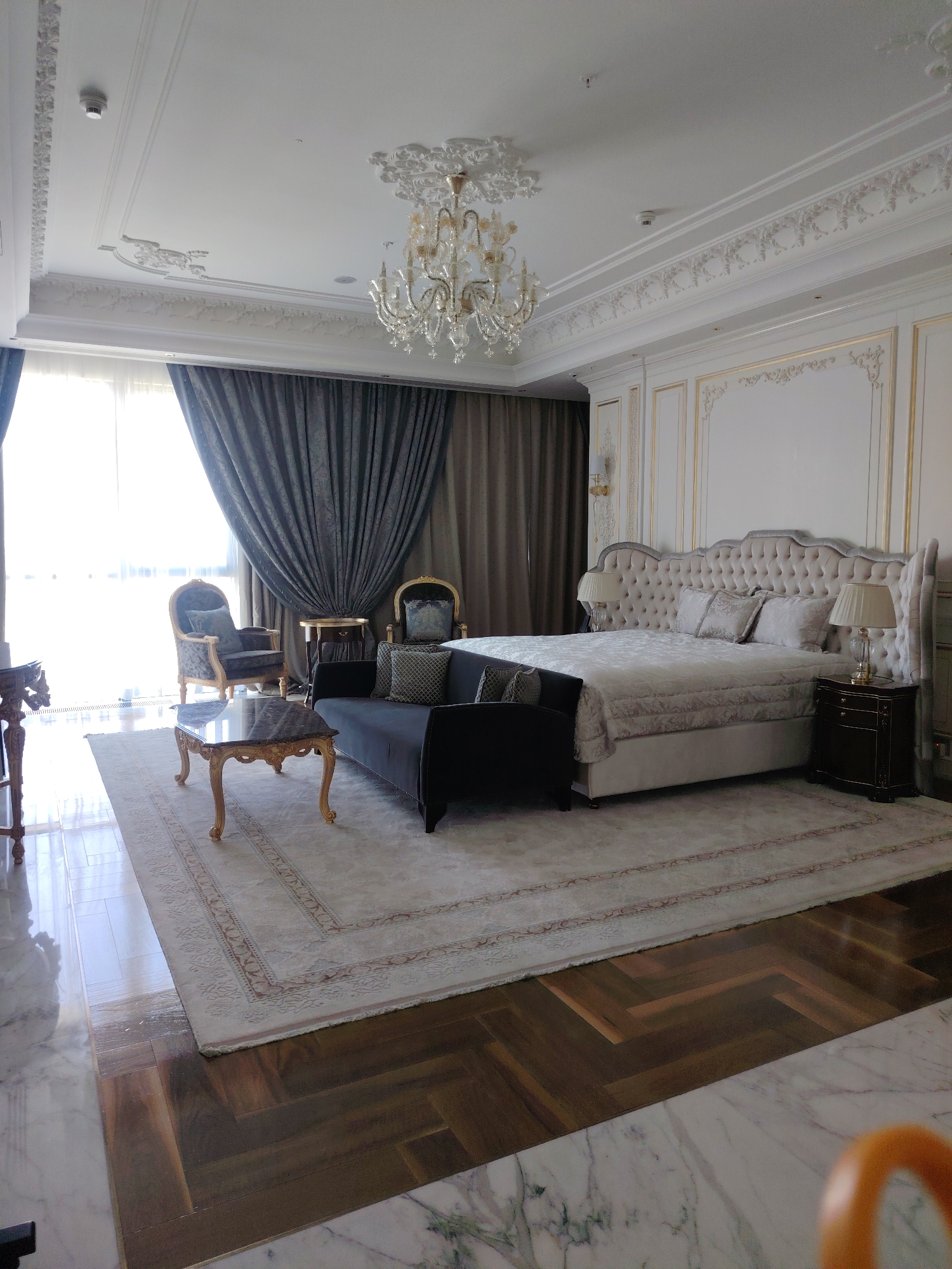 A lavishly decorated bedroom in Konigil, Uzbekistan, features a large, tufted bed with a cream-colored headboard and ornate patterned bedding. Two glass-and-brass bedside lamps flank the bed, placed on dark wood nightstands with ornate detailing. A dark upholstered sofa with decorative pillows sits at the foot of the bed. In the foreground, a small, rectangular coffee table with a marble top and gilded legs rests on a large, cream-colored rug with a patterned border. Two ornate armchairs with dark upholstery and gold accents are positioned near a large window covered by heavy, dark grey curtains. The room has high ceilings with intricate decorative molding and a crystal chandelier. The flooring is a combination of polished marble and dark wood laid in a herringbone pattern. The scene is static and shows no individuals.