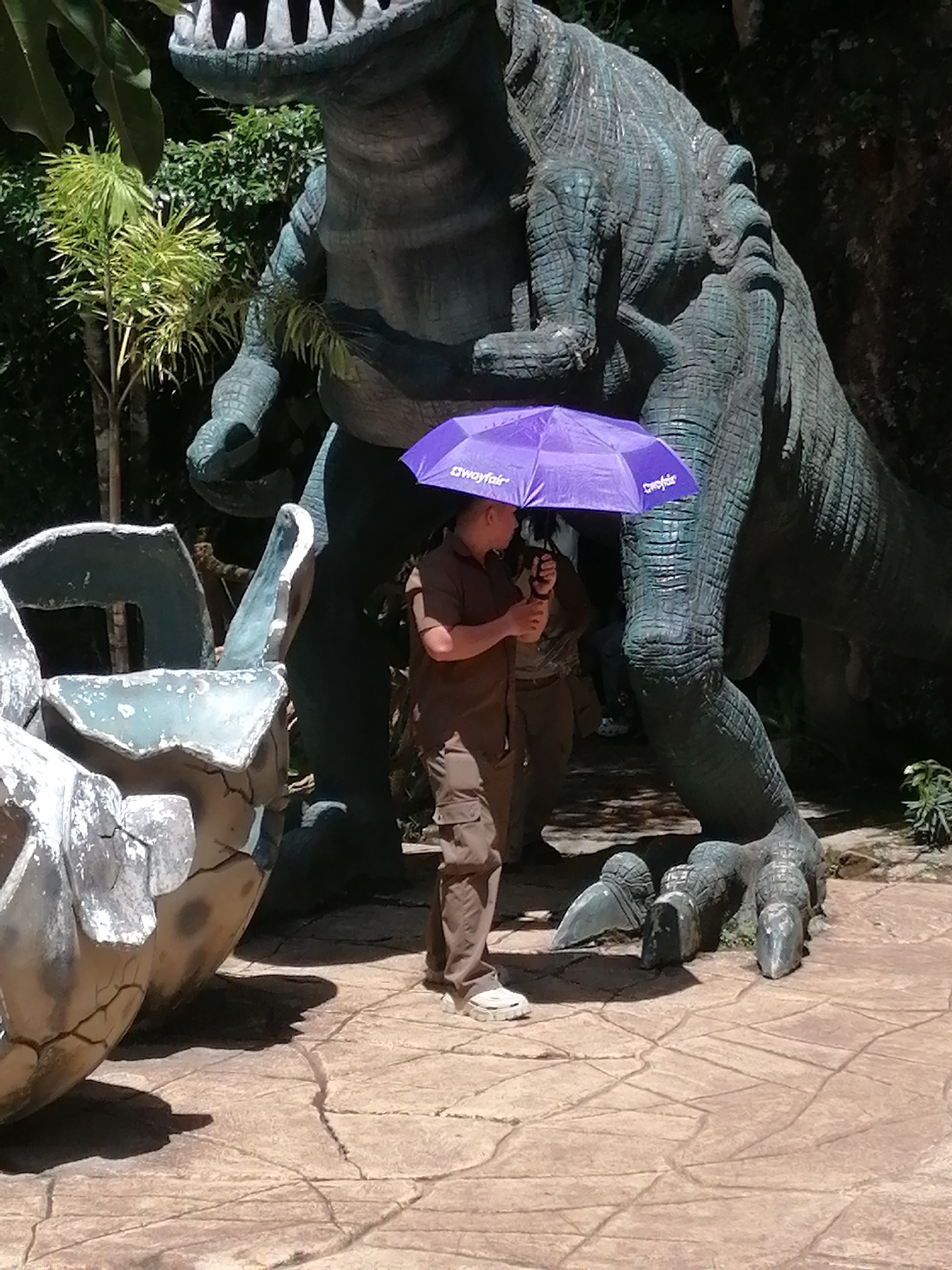 The image depicts a person standing beneath a large, dark green dinosaur statue, holding an open purple umbrella. The dinosaur appears to be a T-Rex, with a massive body, clawed feet, and sharp teeth visible in its open mouth. In the foreground, to the left, is a cracked, egg-like structure, likely part of a display. The ground is paved with textured, light brown stones. Behind the dinosaur, lush green foliage is visible. The scene suggests an outdoor attraction, possibly a theme park or a dinosaur-themed garden. This scene is located at the Dinosaur Park within the Datanla High Rope Course in Da Lat, Vietnam.