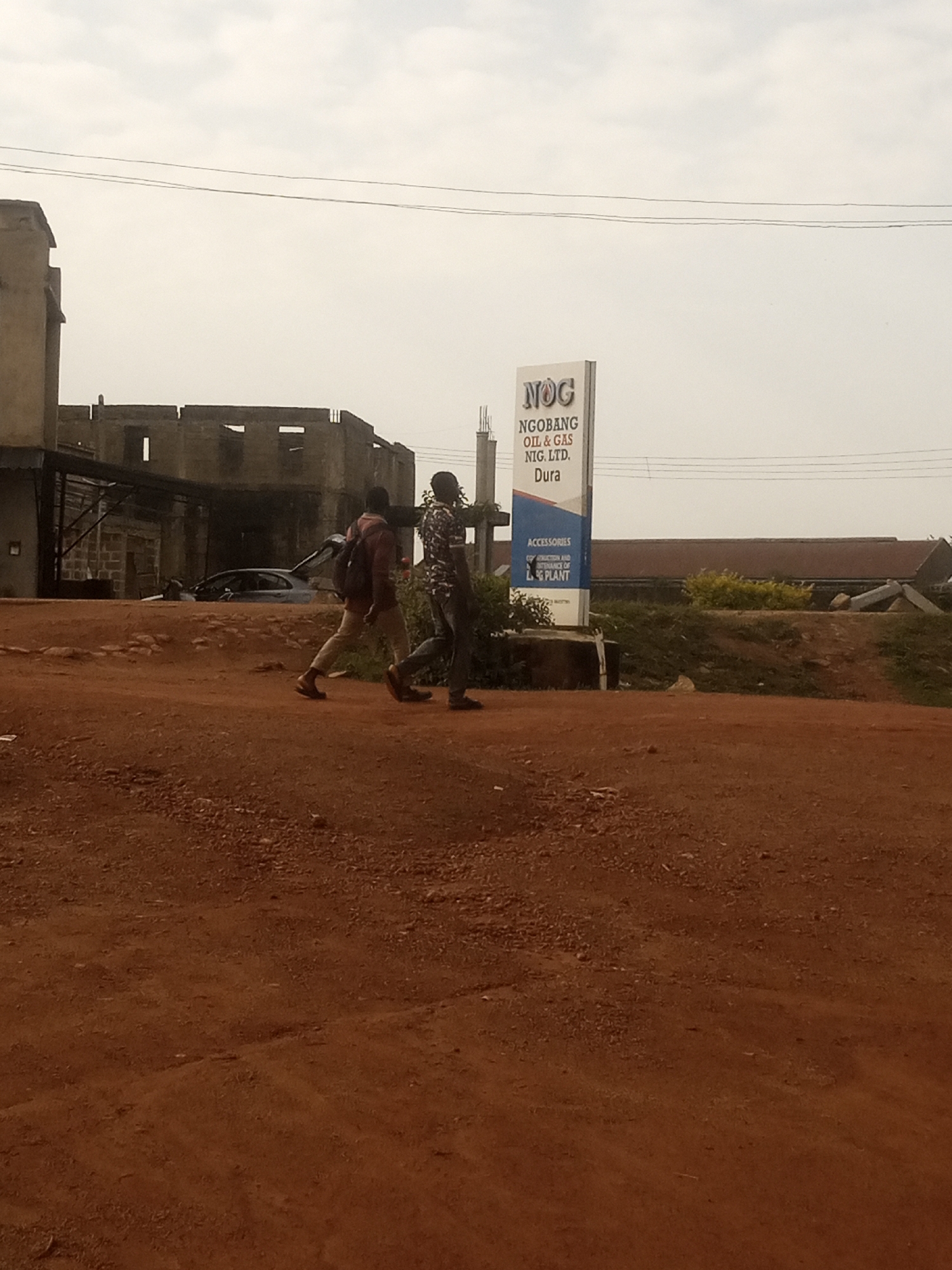The image depicts an outdoor scene in Jos, Nigeria. In the foreground, a reddish-brown dirt path dominates the lower half of the frame. Two individuals are walking across this path from left to right. The person on the left is wearing a dark-colored top and light-colored pants, carrying a backpack. The person on the right is wearing a patterned shirt and dark pants.
In the midground, there is a tall sign with text that appears to read "NOG NGOBANG OIL & GAS NIG. LTD. Dura" and "ACCESSORIES CONSTRUCTION AND MAINTENANCE OF GAS PLANT". Behind the sign, a multi-story building with exposed brickwork and unfinished construction is visible. A car with its trunk open is parked in front of this building. To the right of the sign, there is a grassy embankment with some vegetation.
The background features a hazy sky with thin, horizontal power lines stretching across it. The overall impression is that of a commercial or industrial area in a state of development or disrepair.