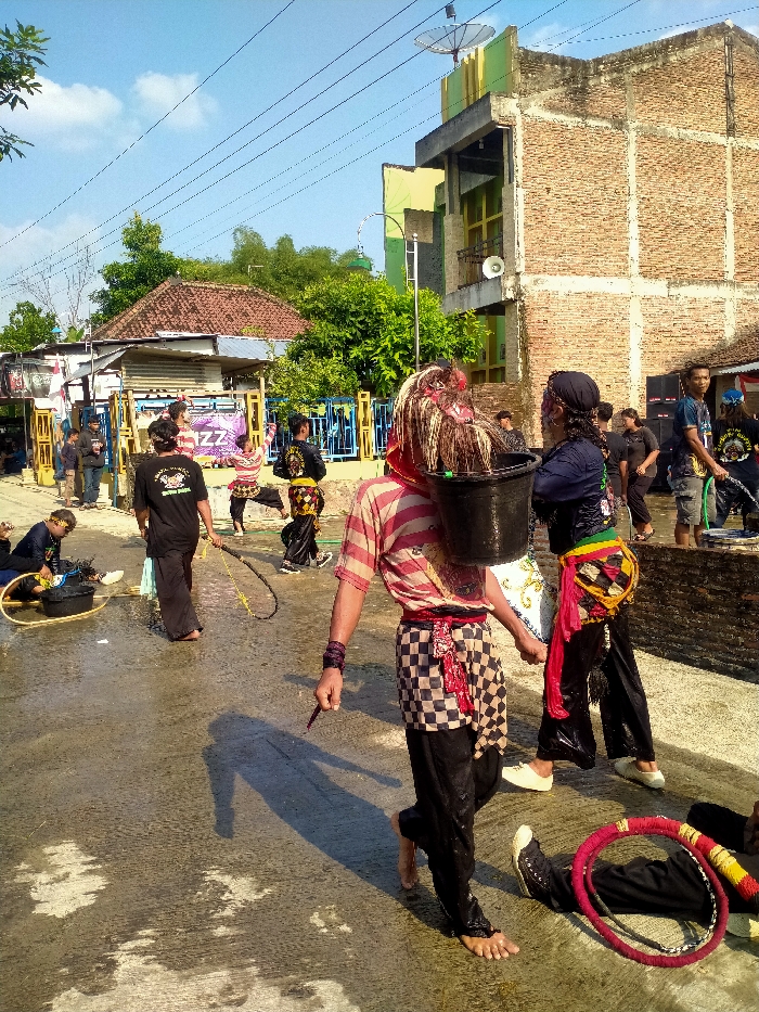The image depicts an outdoor daytime scene on a wet, paved street in Sepat, Indonesia. Approximately fifteen individuals are present, several of whom are dressed in performance or traditional attire.
In the immediate foreground, a barefoot individual is captured mid-stride, moving from right to left. This individual wears a horizontally striped shirt (pink and white), black trousers, and a checkered brown and white skirt-like garment over the trousers, secured by a red sash. A black bucket hangs from their neck, obscuring part of their face, and a light-colored, shaggy headpiece with red elements is visible. A small, dark stick is held in their right hand. Adjacent to this individual, another person stands wearing a dark long-sleeved shirt, black trousers, and multiple colored sashes (red, yellow, green) around their waist.
In the mid-ground, a person in a black shirt holds a long, flexible, black whip-like object. Further left, another individual is seated on the wet ground, attending to a black bucket. Two additional performers, dressed in black attire with yellow or gold embellishments, are captured in a dynamic, bent-knee posture.
On the far right, a person is observed holding a green hose, contributing to the wet state of the pavement. In the lower right corner, a person is lying on the ground next to a large red and black striped circular object, resembling a hoop. Black speakers of a sound system are positioned on the right side near a building.
The background features a multi-story brick building on the right, partially painted light green and yellow, with a satellite dish mounted on its roof. On the left, several single-story residential structures with reddish-brown tile roofs are visible behind a blue fence. A purple banner displaying partial text, including "ZZ," is strung between poles in the mid-background. Numerous overhead power lines crisscross the clear blue sky. The paved ground exhibits extensive wet patches and reflections.