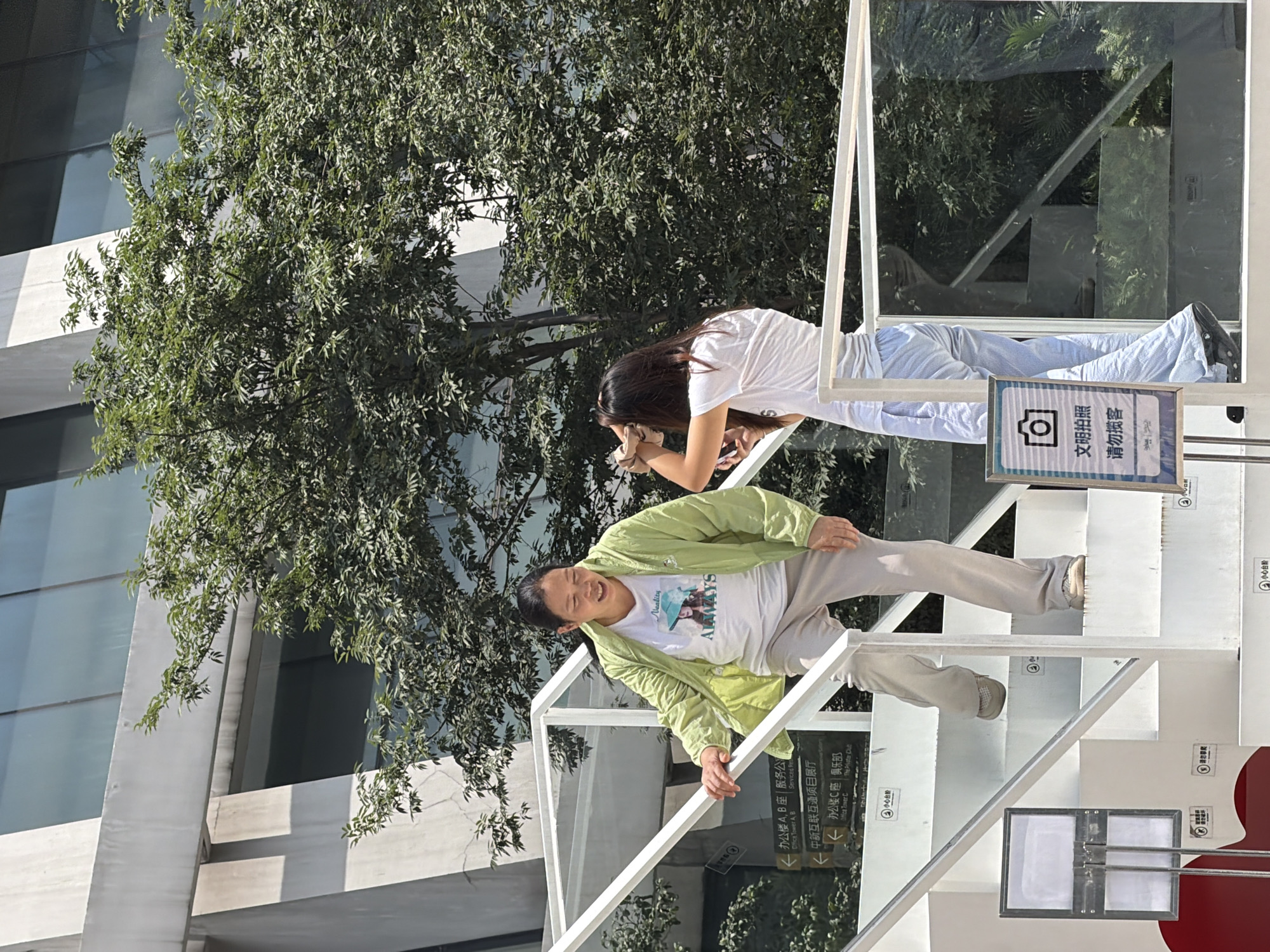 Two individuals are on a white staircase outdoors. The person on the left, a woman with dark hair pulled back, wears a light green jacket over a white t-shirt with a graphic and light-colored pants. She is facing the other person and looking down, smiling. The person on the right, with long dark hair, is wearing a white t-shirt and striped light blue pants. They are holding a phone and a light-colored object near their face, possibly adjusting their hair or taking a picture. The staircase is modern with white treads and rails, some with glass panels. Green trees and a modern building with large windows form the background. Signs with Chinese text are visible on the side of the staircase, one with a camera icon. The setting appears to be an urban area in Yuzhong District, China, on a bright day.