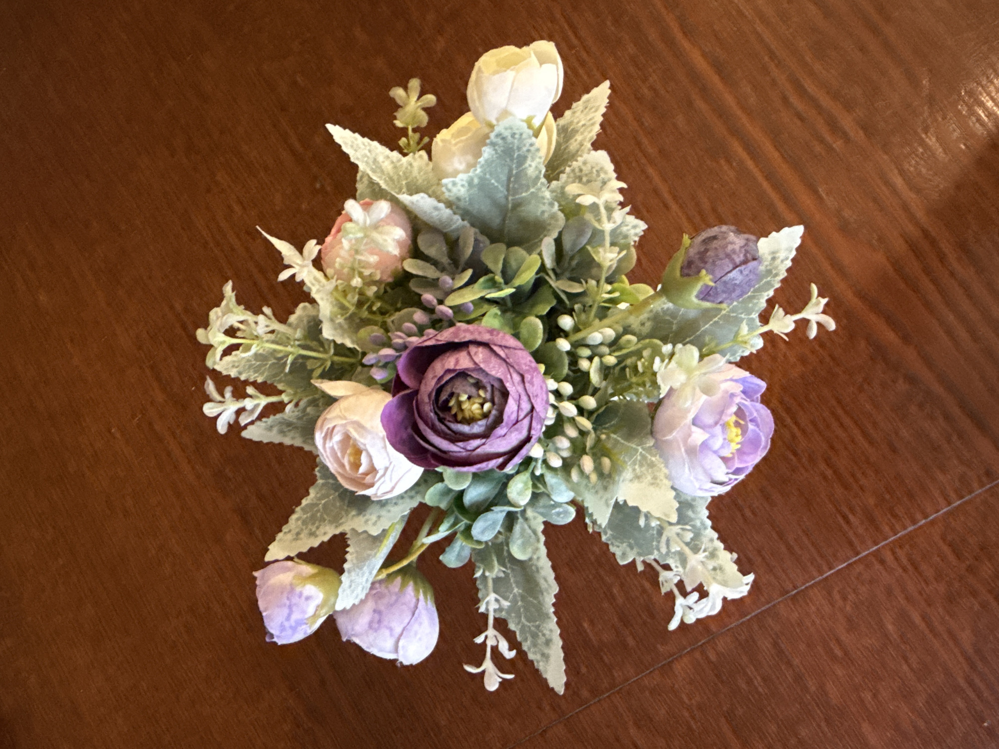 A bouquet of artificial flowers, featuring a prominent deep purple flower at its center, is arranged on a dark brown, polished wooden surface. The bouquet includes several other flowers in shades of light pink, lavender, and cream, along with a variety of green leaves and small white floral accents. The flowers are clustered together, creating a visually appealing arrangement. The wooden surface appears to be a tabletop or desk, with a visible grain pattern and reflections indicating a polished finish. The lighting is soft, casting subtle shadows that highlight the textures of the flowers and the wood. There are no people or animals present in the image.