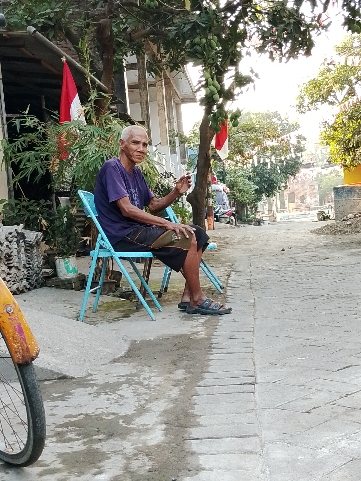 An elderly Indonesian man with short, grey hair and a dark purple t-shirt sits on a light blue folding chair on a paved walkway. He is wearing dark shorts and black sandals. He holds a cigarette in his right hand, which is raised. The man is looking towards the left of the frame. Behind him, a red and white Indonesian flag is visible, attached to a bamboo stalk. Trees with green foliage and what appear to be unripe mangoes hang in the background. The walkway is made of grey bricks and has wet patches on it, suggesting recent rain. To the left of the man, a pile of stacked grey tiles is visible. In the foreground, part of a bicycle wheel with an orange and yellow fender is visible on the left side of the frame. The scene appears to be in an outdoor, possibly residential area, with buildings and more trees in the distance. The presence of the Indonesian flag suggests the location is in Indonesia. Given the context of the city of Pasuruan, this scene likely depicts a moment of rest or daily life in one of its neighborhoods.