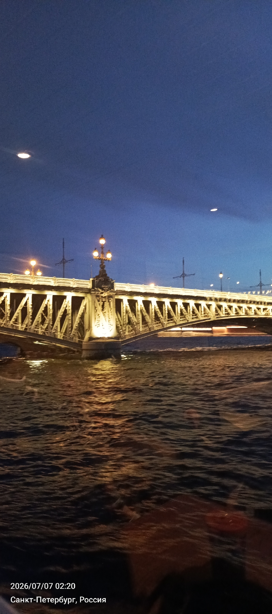 The image captures a nighttime view of a bridge spanning a body of water in Saint Petersburg, Russia. The bridge is illuminated by streetlights, with the warm glow reflecting on the rippling water below. The sky is a deep blue, suggesting either dusk or dawn. The architecture of the bridge is intricate, featuring a lattice-like metal structure and decorative lampposts. The water appears somewhat choppy, indicating a current or gentle waves. The timestamp in the lower left corner reads "2026/07/07 02:20" and the location is identified as "Санкт-Петербург, Россия" (Saint Petersburg, Russia). No people are clearly visible in the image, though there are dark areas at the bottom that might suggest the presence of observers. The overall scene is serene and atmospheric, highlighting the urban landscape at night.