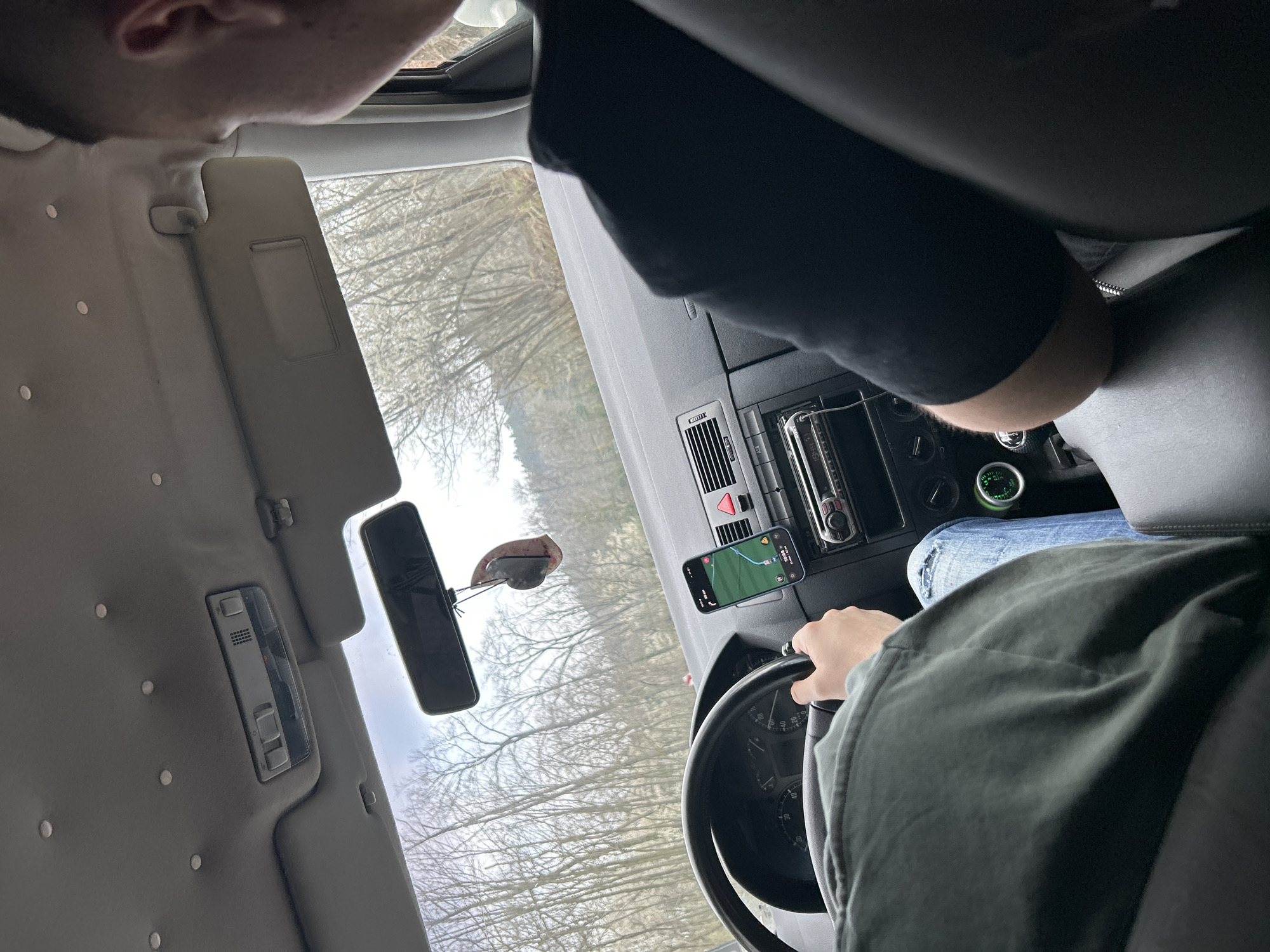 A view from inside a car showing the driver holding the steering wheel, a smartphone mounted on the dashboard displaying a map application, and a glimpse of trees through the windshield. The car's interior is visible, including the dashboard, radio, rearview mirror, and sun visor. The driver is partially visible, wearing a light blue jeans.
Location: Unknown, Unknown.