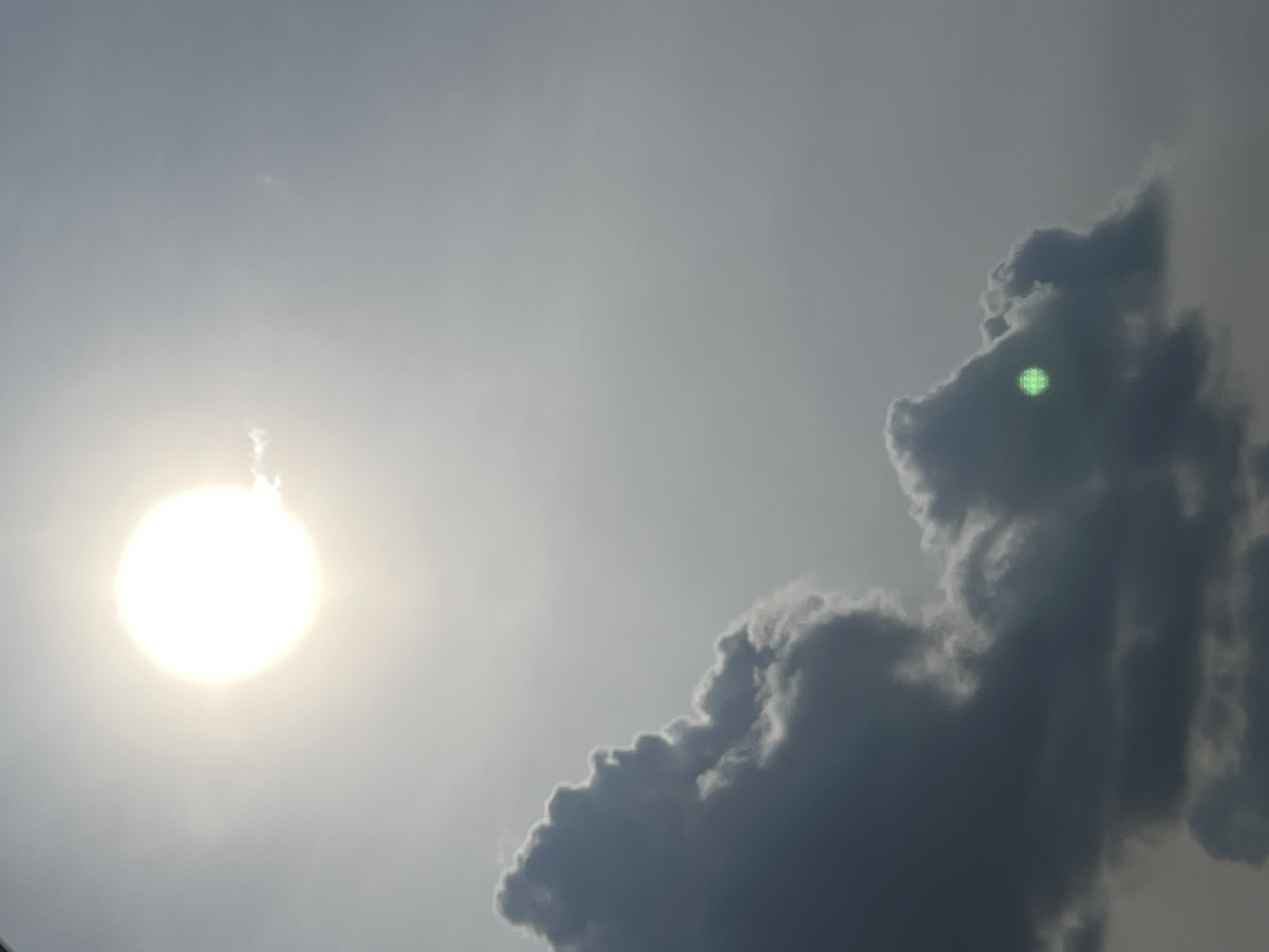 The image captures a bright, hazy sun partially obscured by dark, dramatic clouds against a muted gray sky. The sun, located in the upper left quadrant of the frame, appears as a large, luminous orb with a diffused halo, suggesting it might be viewed through atmospheric haze or a thin layer of clouds. In the lower part of the image, substantial dark clouds dominate, with their edges softly illuminated by the sunlight. One prominent cloud formation in the lower left has a sharp, angular profile, while another in the lower right is more rounded and billowy. A small, distinct green light, possibly a lens flare or an artifact, is visible near the center of the right cloud formation. The overall scene suggests a dynamic weather condition, possibly before or during a storm, with contrasting light and shadow. The image does not contain any identifiable landmarks or elements that would indicate its specific location within Warsaw, Poland.
