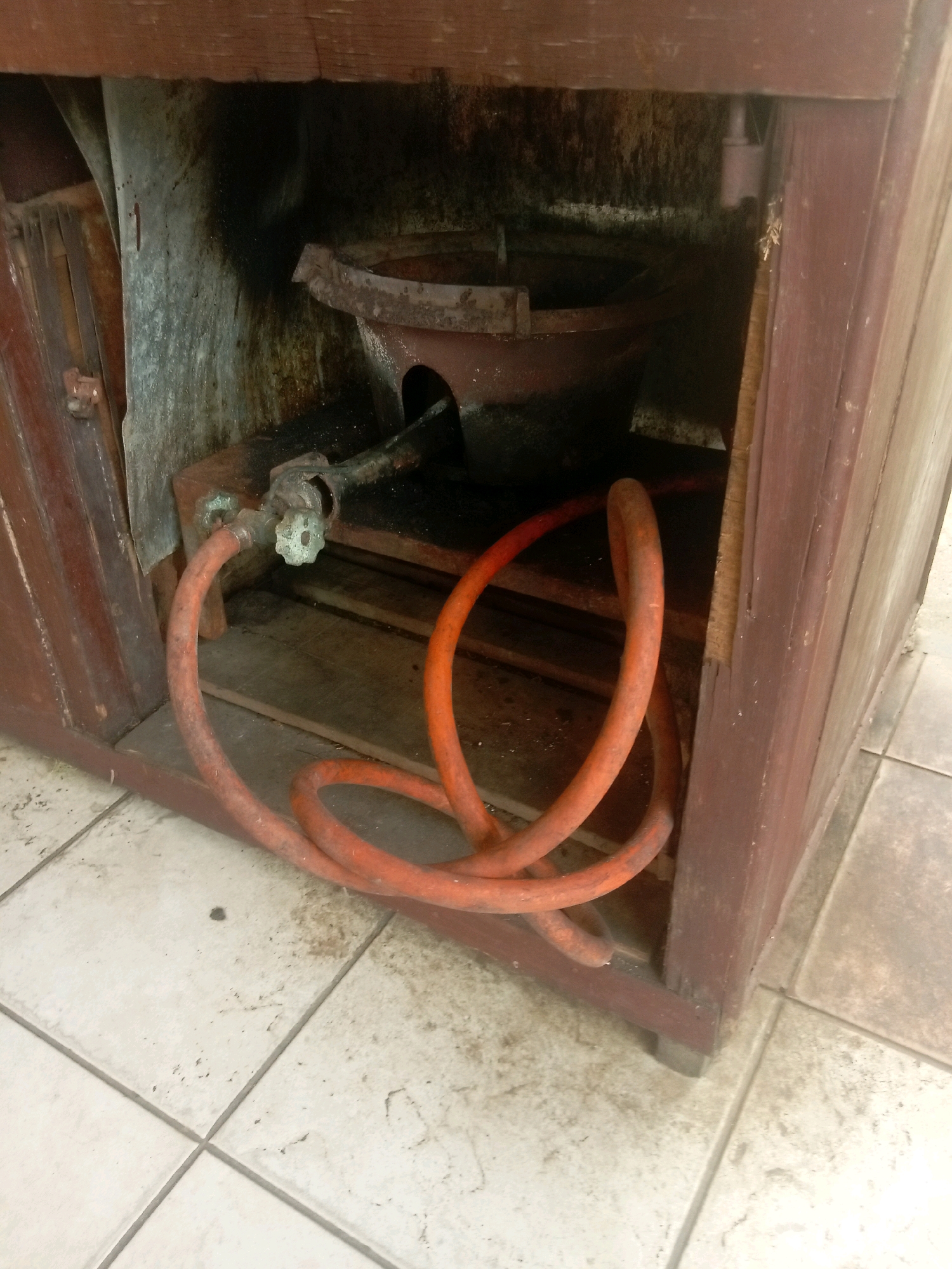 An outdoor view of a gas stove integrated into a wooden cabinet with a visible gas hose running from a valve to the stove. The scene is set on tiled flooring, typical of outdoor spaces or kitchens in Batam, Indonesia. The stove appears old and somewhat dirty, with soot stains visible on the interior walls of the cabinet and around the stove itself. The gas hose is bright orange and coiled loosely on the floor in front of the cabinet. The wooden cabinet is aged and shows signs of wear. The overall impression is one of an older, possibly infrequently used, cooking setup in an outdoor or semi-outdoor environment.