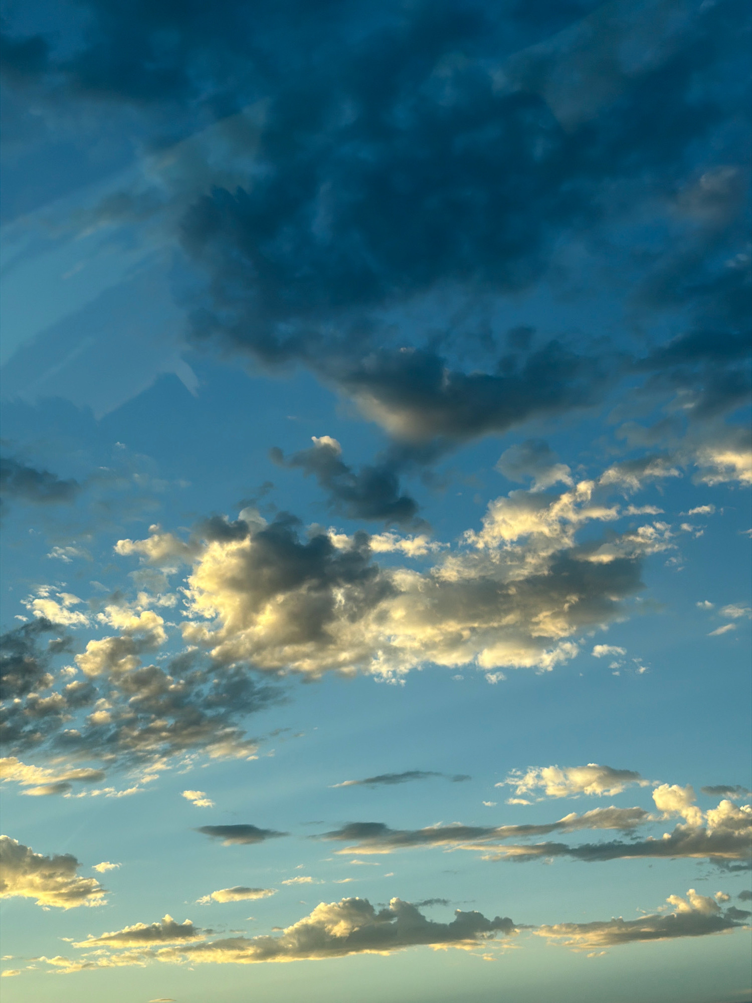 The image shows a sky filled with clouds. The clouds are varying in color from dark blue to white and yellow, indicating different lighting conditions and cloud densities. Some clouds are large and appear to be storm clouds, while others are smaller and wispy. The sky itself is a gradient of blue, with lighter shades near the horizon and darker shades towards the top. There is no discernible city or specific location visible in the image.