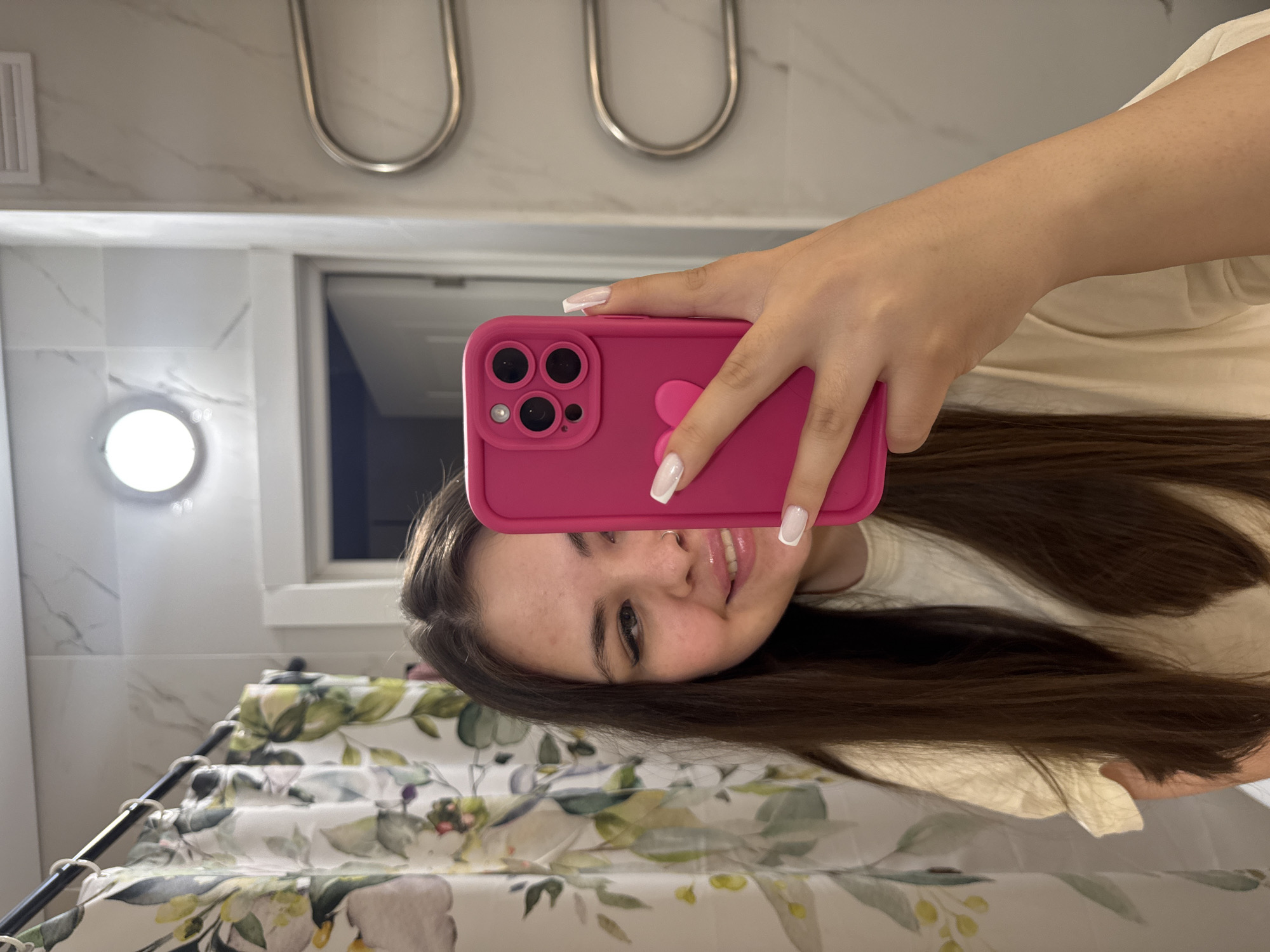 A young woman is taking a selfie in a bathroom. She is holding a pink phone with a protective case. The bathroom has a shower curtain with a floral pattern, white marble-like tiles, and two towel racks. A round light fixture is visible on the wall. The location is likely Murino, Russia, as requested.
