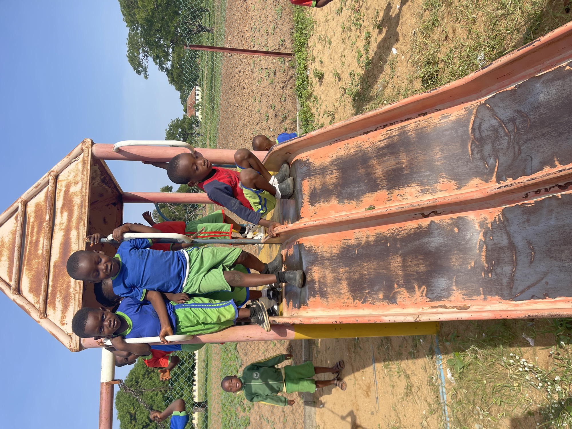 In Akwanga, Nigeria, a group of children plays on an old metal playground slide in an outdoor area. Several children, dressed in various colored shirts and green shorts, are sitting or standing on the slide's platform. One child is standing near the base of the slide in a green outfit. The slide is rusted and worn, and the surrounding area is a mix of grass, dirt, and some vegetation. A chain-link fence separates the playground from a field or open area. The sky is clear and blue.