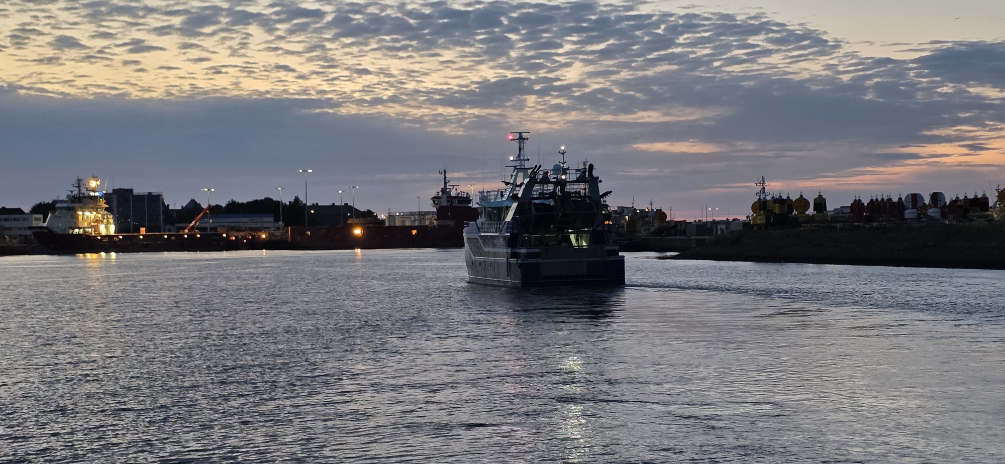 The image shows a harbor scene at dusk in Den Helder, Netherlands. The sky is filled with dramatic clouds, tinged with the orange and pink hues of sunset. In the foreground, the dark water of the harbor reflects the twilight sky and the lights of the surrounding city.
Dominating the center of the frame is a large fishing vessel moving away from the camera. Its stern is visible, showing a dark hull and a lit deck. To the left, another vessel is docked, its lights illuminating the water. In the background, the silhouette of the Den Helder skyline can be seen, with various buildings and industrial structures. The harbor is equipped with lighting poles, and there are also various buoys and other nautical equipment visible on the shore. The overall atmosphere is one of a working harbor winding down for the night.