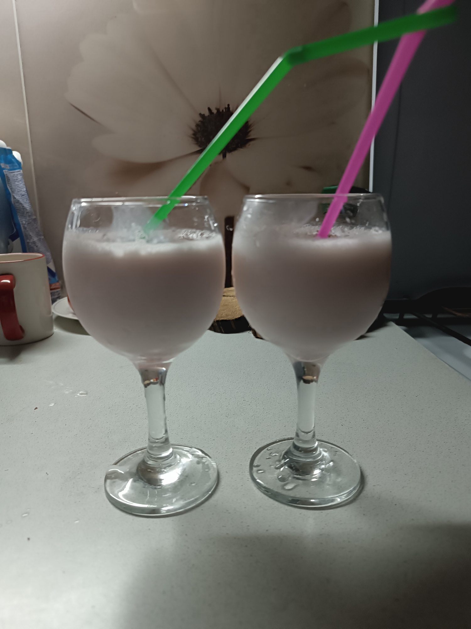 Two wine glasses filled with a pale pink liquid are placed on a light grey countertop. The glass on the left has a green straw, while the one on the right has a pink straw. Behind the glasses is a floral print on a surface. A mug with a red handle is partially visible to the left of the left glass. The setting appears to be indoors, potentially a kitchen, in an unspecified location in the city of Unknown, Unknown.