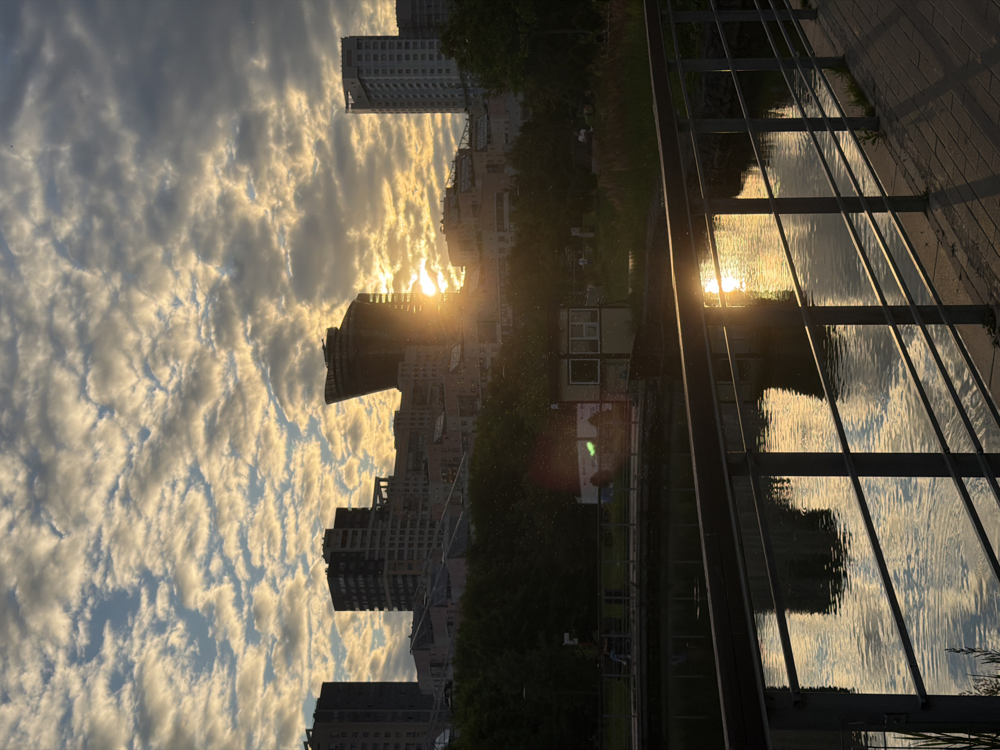 The image shows a city skyline at sunset. The sun is setting behind a cluster of modern buildings, casting a warm glow on the clouds and reflecting on the water in the foreground. The foreground features a body of water with a bridge or walkway railing. The railing is made of metal and wood, and it is positioned in front of the water. The water is calm and reflects the sky and the surrounding buildings. The buildings in the background are tall and modern, with a variety of architectural styles. They are set against a backdrop of a cloudy sky. The overall scene is serene and picturesque, with the sunset creating a beautiful atmosphere.