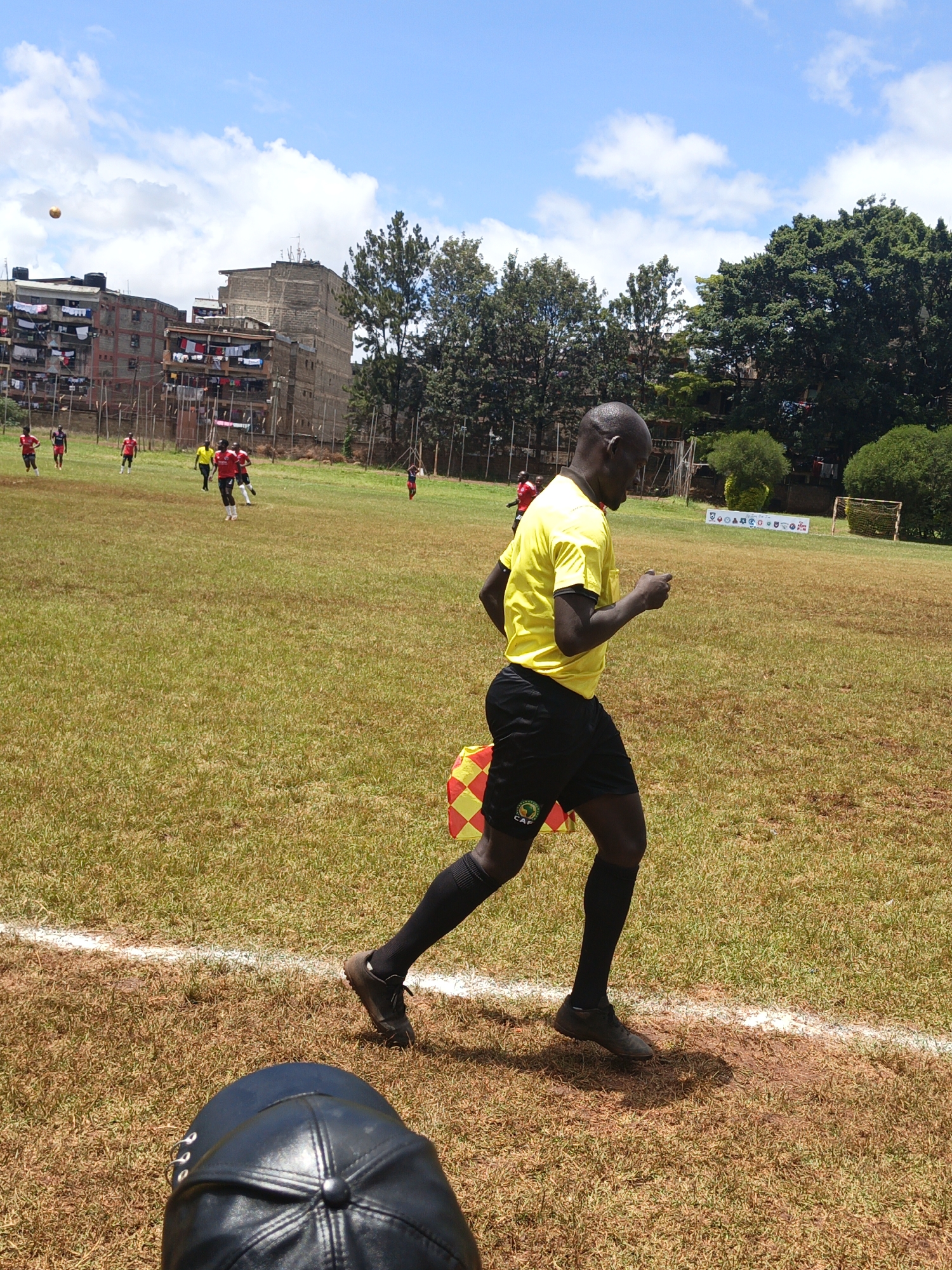 The image captures a soccer match in progress. An assistant referee in a yellow shirt and black shorts runs along the touchline, holding a checkered flag. The players are of African descent. A ball is in the air. In the background, buildings with laundry hanging on the balconies of the city of Nairobi, Kenya, are visible. The field appears to be well-worn with brown and green grass. A soccer goal is visible in the distance. A fence surrounds the field. A black hat is visible in the foreground.