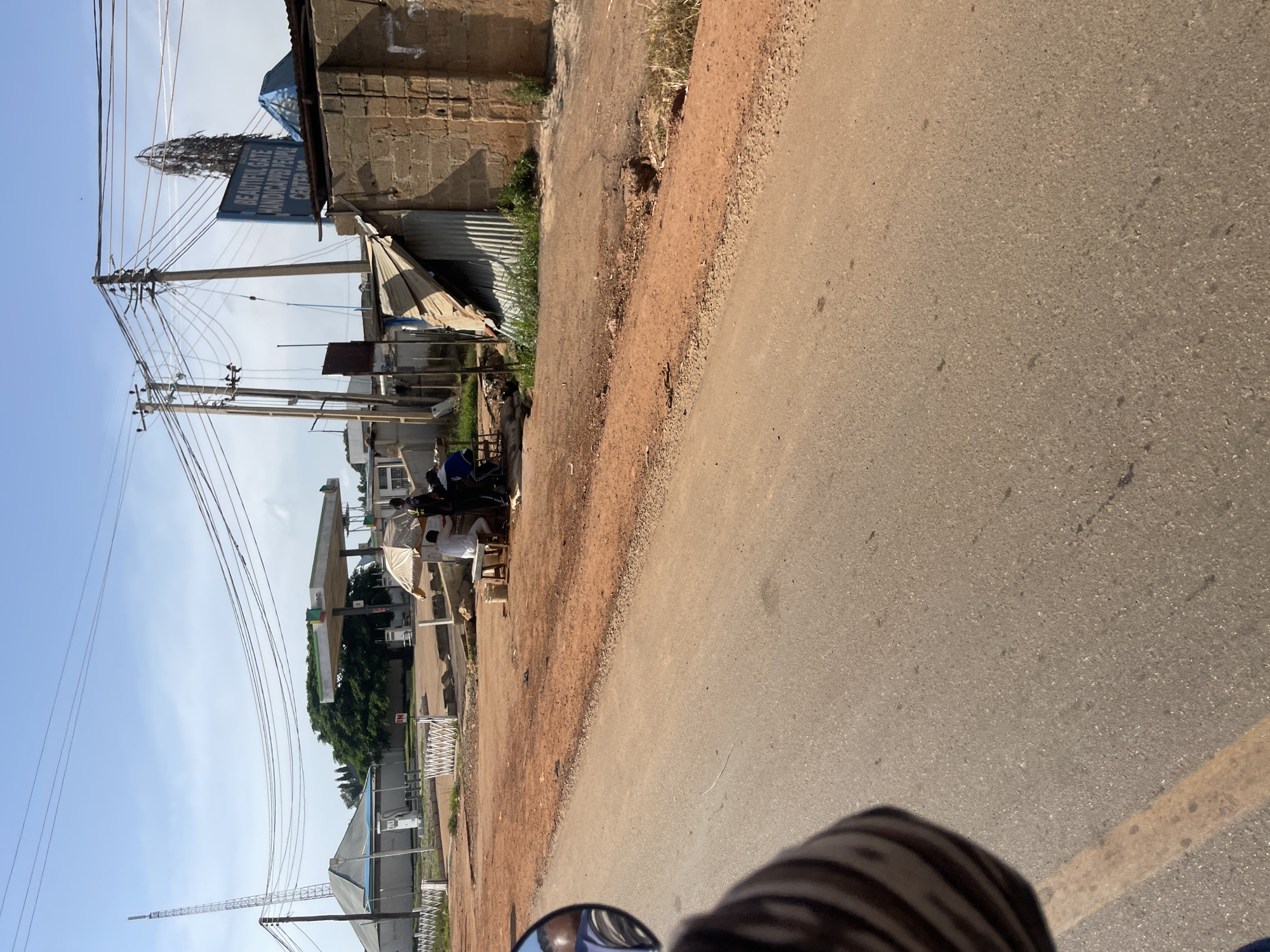 The image shows a street scene in Rafin Par, Nigeria. In the foreground, a portion of a vehicle's side mirror and a person's arm wearing a striped sleeve are visible. The road is asphalt with a yellow lane marking. To the right of the road, there is a red dirt embankment. Further back, a group of people are gathered under a thatched umbrella near a small kiosk. One person is seated on a stool, and another stands behind them. A gas station with pumps and a canopy is visible in the background, along with a tall radio tower and some buildings. A blue sign that reads "BEAUTIFUL GATE HANDICAPPED PEOPLE CENTER" is mounted on a pole. The sky is clear and blue with some clouds. The environment appears to be a busy commercial area with elements of infrastructure and local commerce.