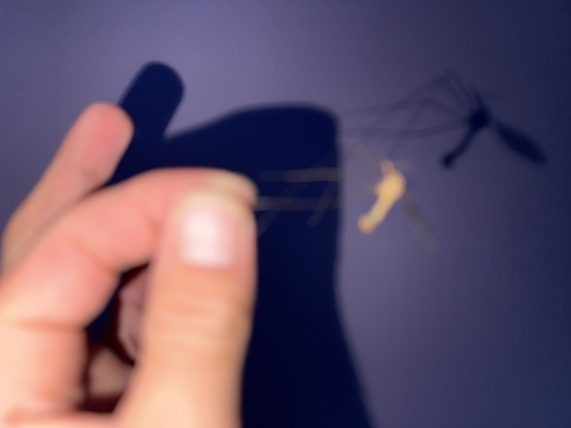 A person's hand, with visible skin texture and fingernails, is blurred in the foreground. The fingers are positioned as if holding or manipulating something thin and delicate. A dark shadow is cast behind the hand, suggesting a light source from the front. In the middle ground, a small, yellow, insect-like object with long, thin appendages is suspended by nearly invisible threads. This object is also out of focus. The background is a solid, dark purple surface, possibly a wall or fabric. There are no discernible urban features or specific locations within a city.