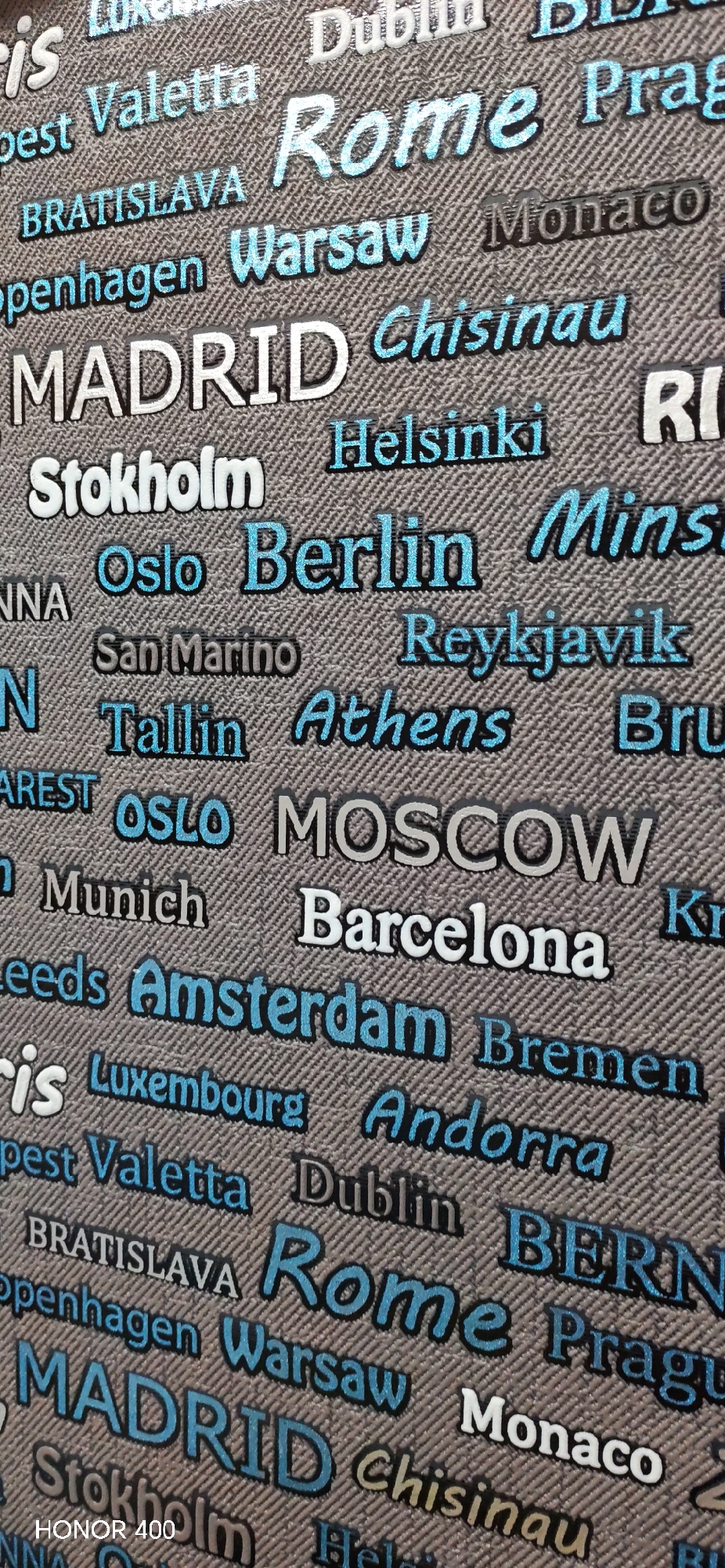 This close-up image displays a patterned textile surface, likely fabric or wallpaper, adorned with numerous names of cities, predominantly European capitals. The background features a subtle, diagonal woven texture in a neutral grey hue.
The city names are rendered in diverse styles, creating a dynamic visual effect. Some names are depicted in a bold, white, all-caps font (e.g., MADRID, BRATISLAVA, OSLO), suggesting an embossed or raised texture. Others are in a vibrant, light blue glitter effect, often italicized (e.g., Rome, Chisinau, Reykjavik, Andorra) or in a block font (e.g., Warsaw, Helsinki, Berlin, Amsterdam). A third style uses a muted grey color, sometimes in all caps (e.g., MOSCOW) or title case (e.g., Monaco, San Marino, Munich, Bremen). The pattern appears to repeat across the surface.
Visible city names include Valletta, Rome, BRATISLAVA, Warsaw, Monaco, MADRID, Chisinau, Helsinki, Stockholm, Oslo, Berlin, San Marino, Reykjavik, Tallinn, Athens, MOSCOW, Munich, Barcelona, Amsterdam, Luxembourg, Andorra, Dublin, and Bremen, alongside partial views of others like Prague, Copenhagen, Minsk, Brussels, Bucharest, Leeds, and BERN.
No specific activity or event is taking place; the image captures a static pattern. There are no cues for time of day, weather, or emotions. In the bottom left corner, the text "HONOR 400" is visible, likely indicating the device used to capture the image. While the provided location context is Noviy, Russia, the content of the image itself does not depict this location but rather a collection of city names as a design element.
