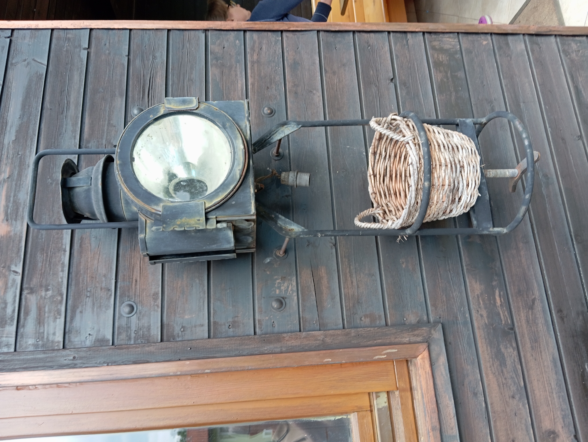 An old, dark wooden shed wall is shown, with a vintage train lamp attached. The lamp has a large, round glass lens and a dark metal casing. Below the lamp, a wicker basket is suspended from a metal frame. A window with a light brown wooden frame is visible on the left side of the image. To the right, a doorway leads into another part of the building, where a person wearing glasses and a dark blue top is partially visible, along with a wooden bench. The image is taken in Baden, Austria.