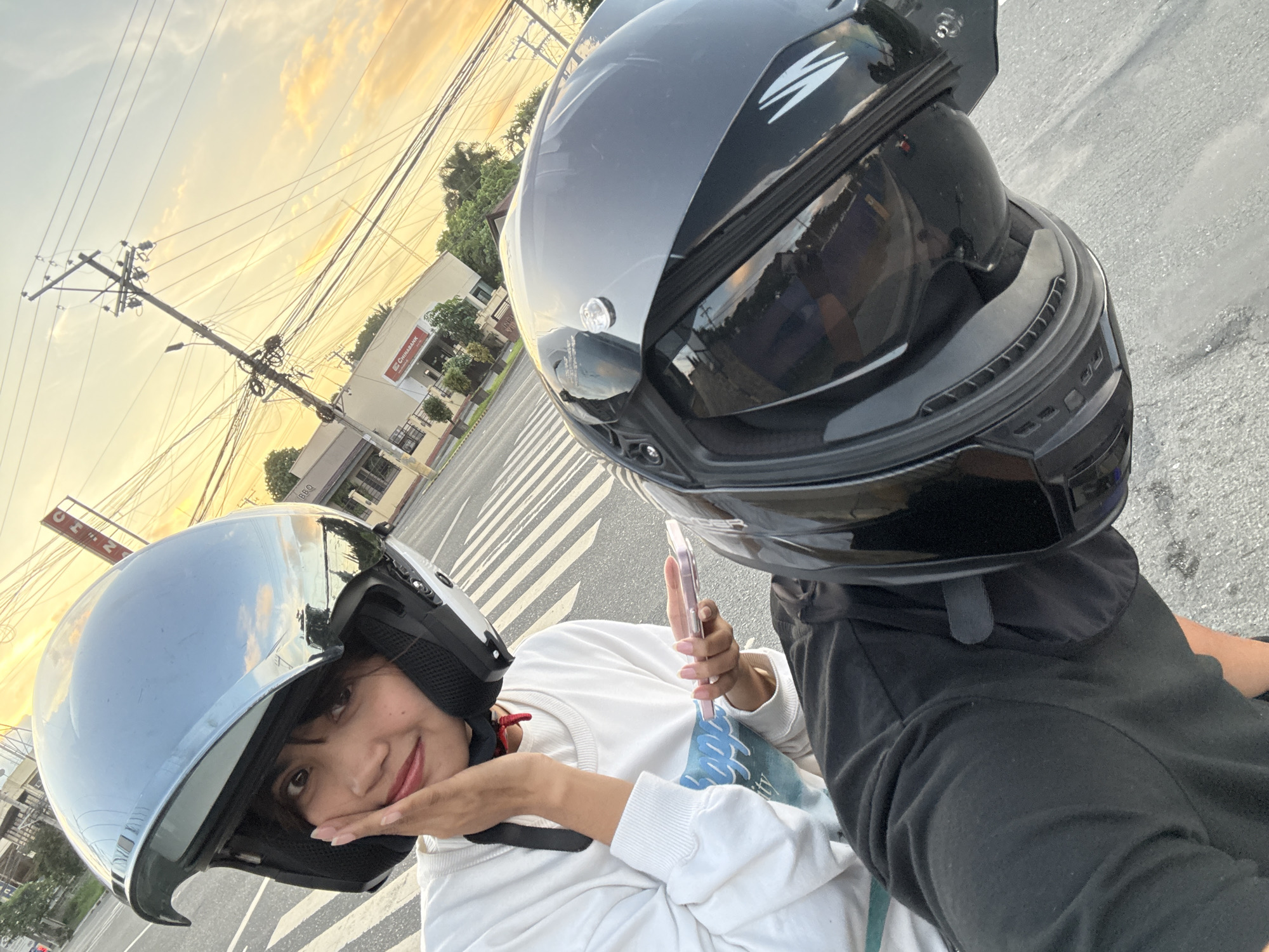 A young woman wearing a silver open-face helmet and a white long-sleeved shirt is captured in a selfie, resting her cheek on her hand. She holds a pink smartphone in her other hand. To her right, another person wearing a black full-face helmet, only partially visible, appears to be driving. The duo is on a street with a crosswalk, and buildings, including a "Chinabank" branch, and a utility pole with tangled wires are visible in the background. The sky displays an orange and yellow sunset. The scene is identified as being in Davao City, Philippines.